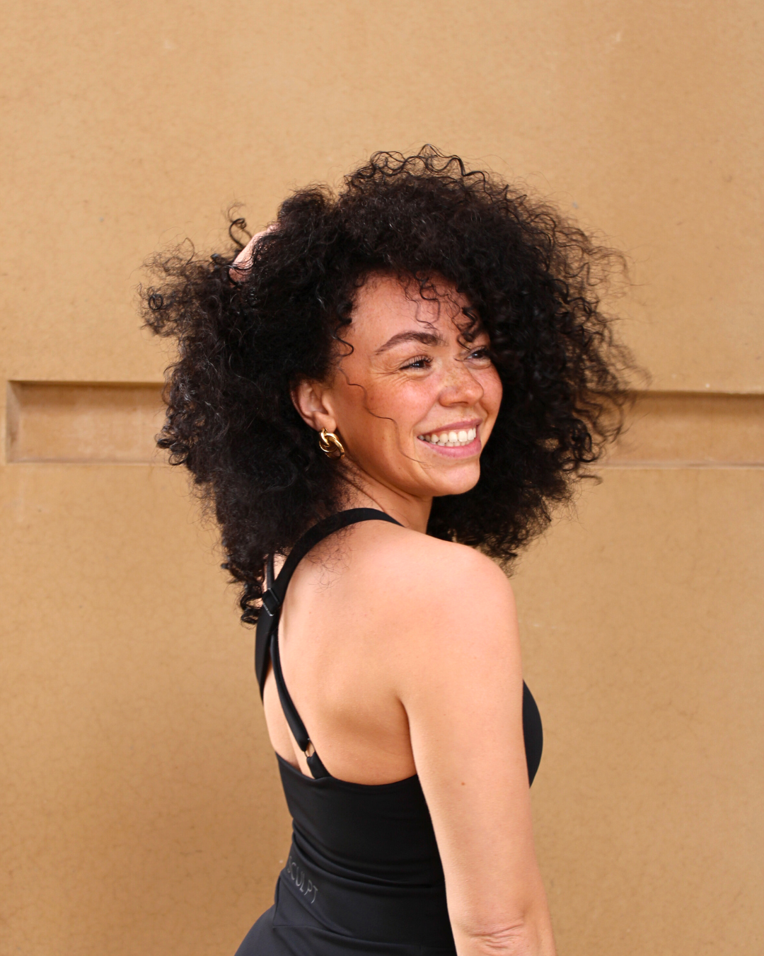 A woman with curly black hair smiling and looking over her shoulder, wearing a black tank top and gold earrings.