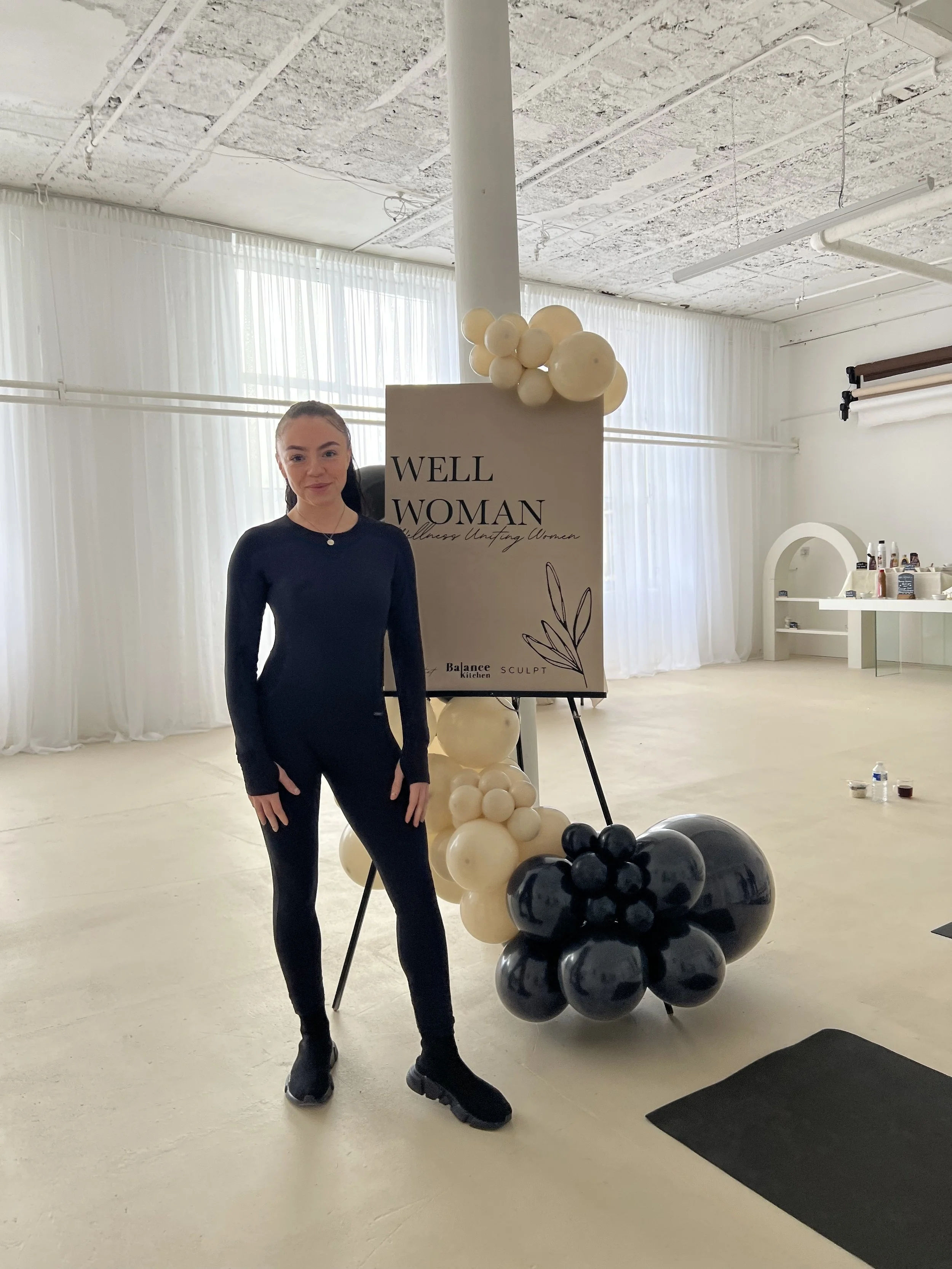A young woman in black athletic wear smiling in a minimalistic, well-lit studio with white sheer curtains. Behind her is a sign that says "WELL WOMAN" and an arrangement of black and white balloons.