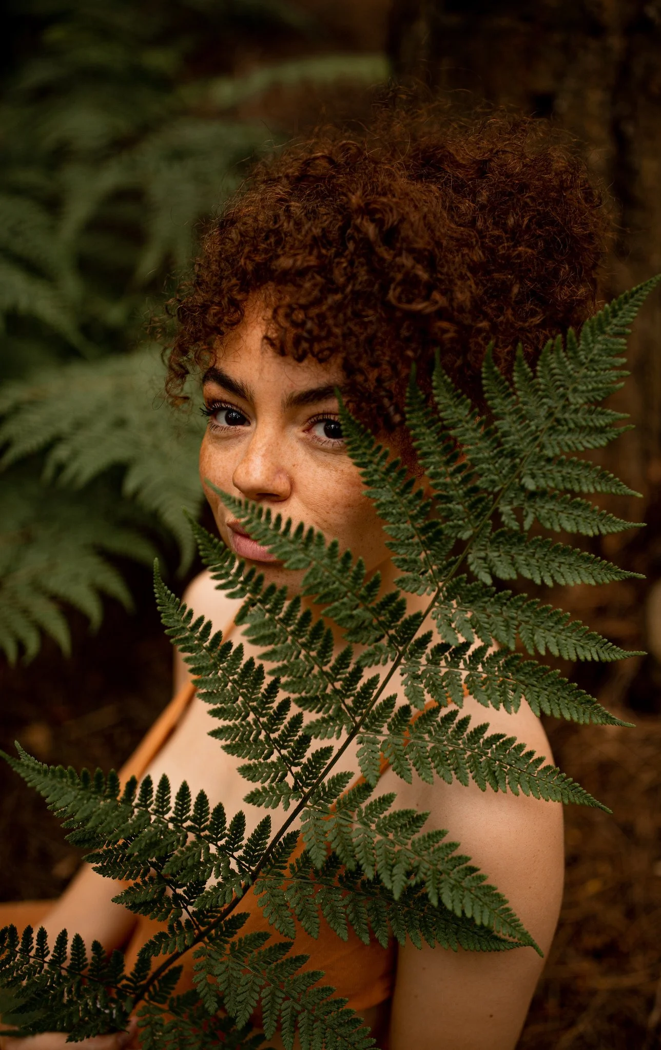 Young woman with curly hair partially hidden behind fern leaves in a forest setting.