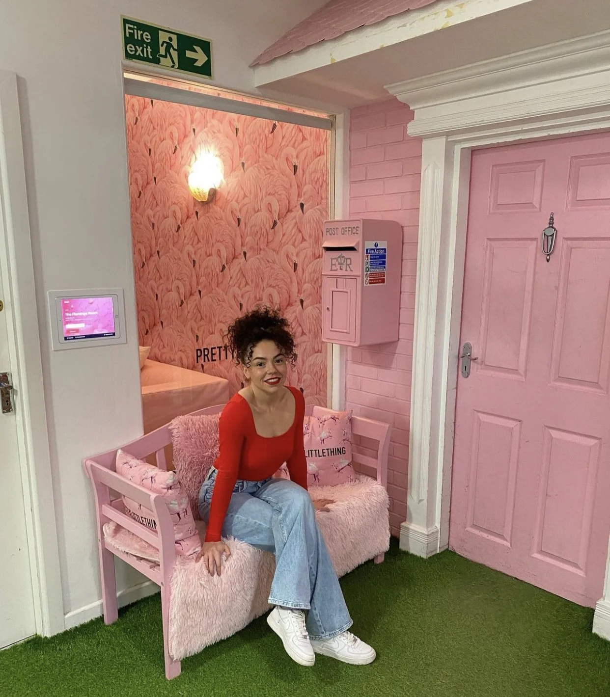 A woman with curly hair wearing a red top, light blue jeans, and white sneakers sitting on a pink furry bench in a pink-themed room with flamingo wallpaper, pillows, and pink doors.