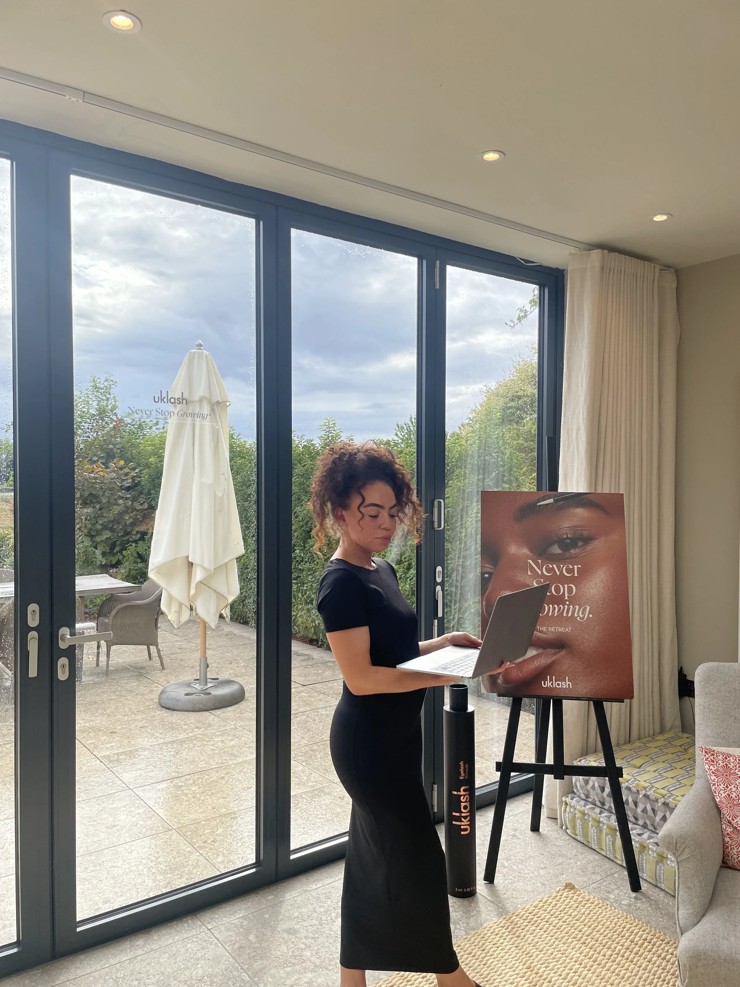 A woman with curly hair stands inside near a glass door, looking at her laptop. There is a large poster with a woman's face and the words 'Never Stop Growing' on an easel nearby. Outside, patio furniture and patio umbrella are visible, and the sky is cloudy.