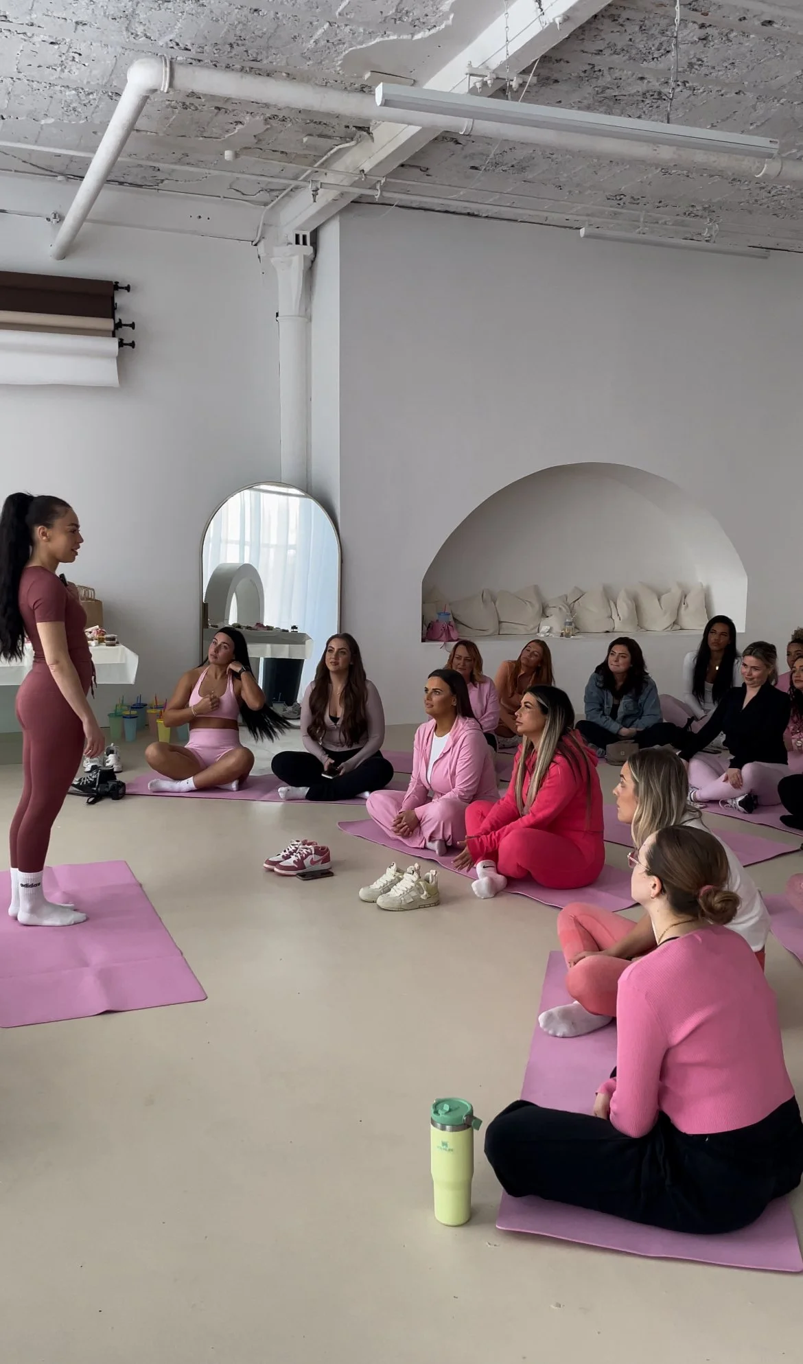 A group of women sitting on pink yoga mats in a dance studio, listening to a woman in pink workout clothes standing at the front, likely giving a fitness or wellness class.