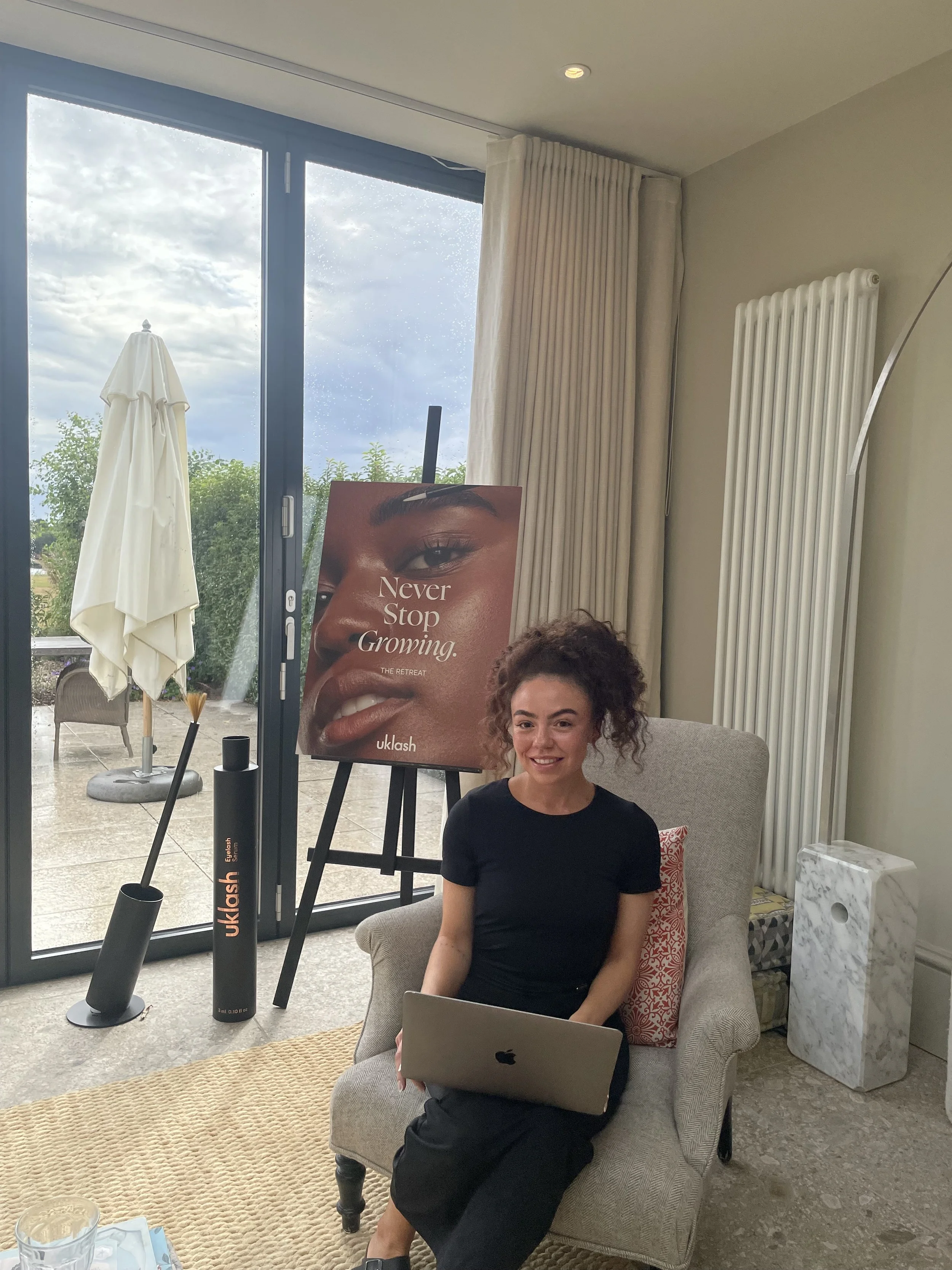 A woman sitting in a beige armchair with a laptop on her lap, smiling, in a room with large windows, a poster of a woman’s face, and a white radiator.