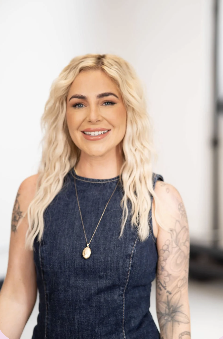 A smiling woman with long, wavy blonde hair, wearing a sleeveless denim dress and a gold necklace, standing in a bright indoor setting.
