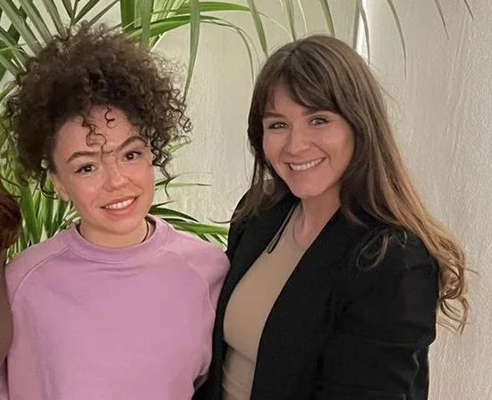 Two women standing next to each other smiling in front of a white wall with green plants.