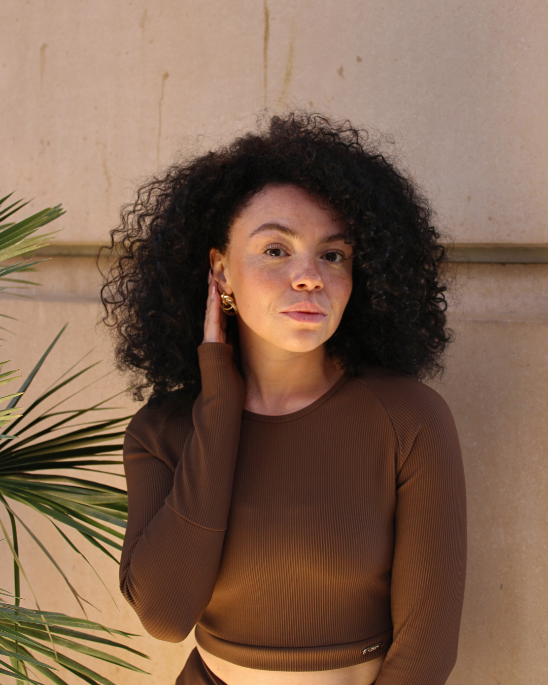 A woman with dark curly hair wearing a brown long-sleeve top standing against a beige wall, with a large green plant nearby.