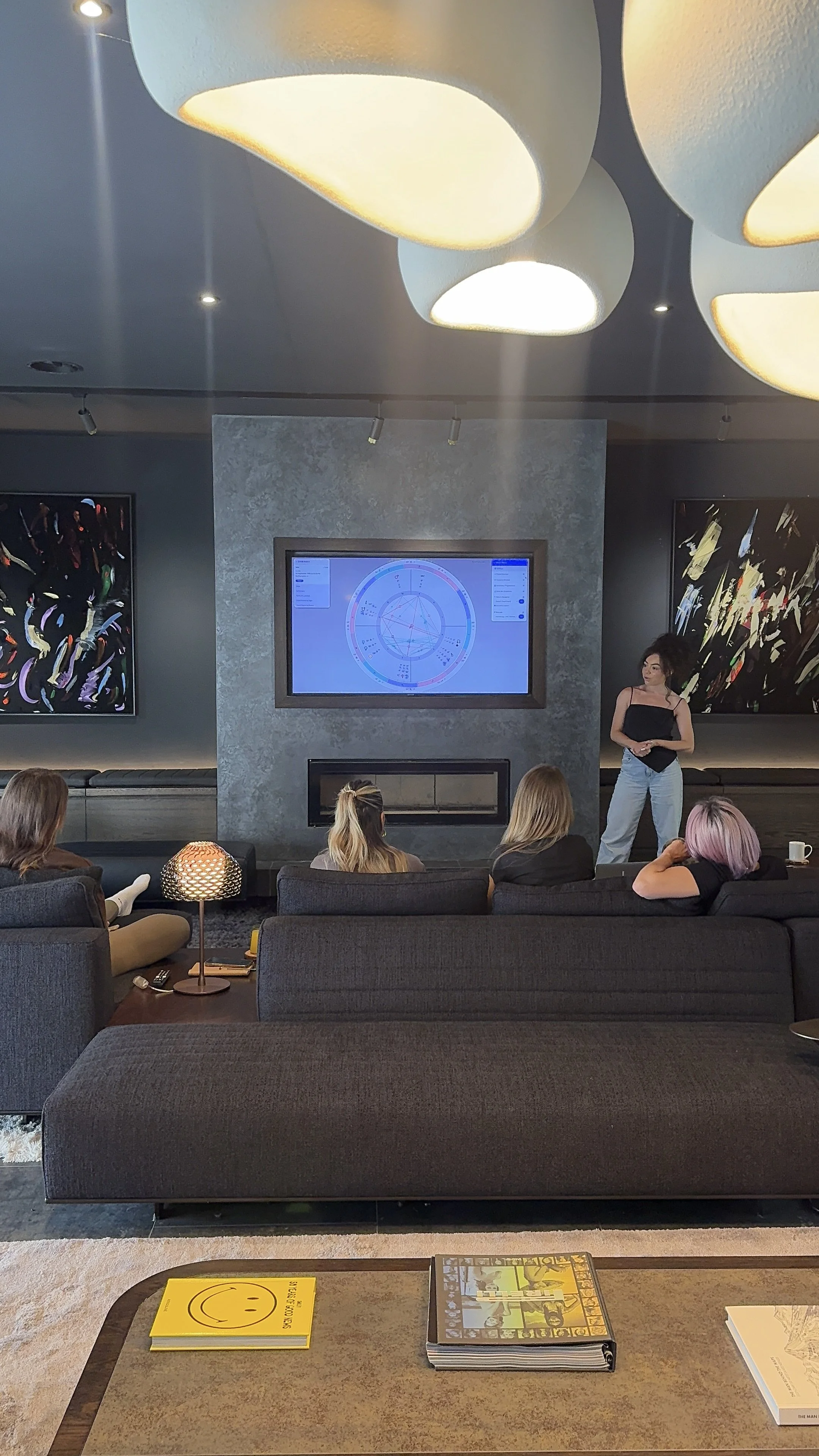 A group of people sitting on gray couches in a modern lounge or conference room, listening to a woman standing near a TV screen displaying a chart or diagram. Modern art on the walls, lamps, and a table with books and a yellow smiley face notebook.
