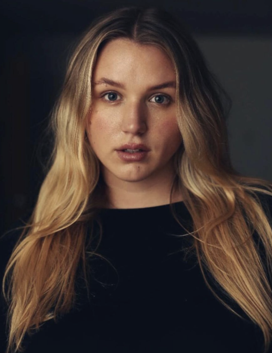 A portrait of a young woman with long, wavy blonde hair, wearing a black top, looking directly at the camera with a neutral expression.