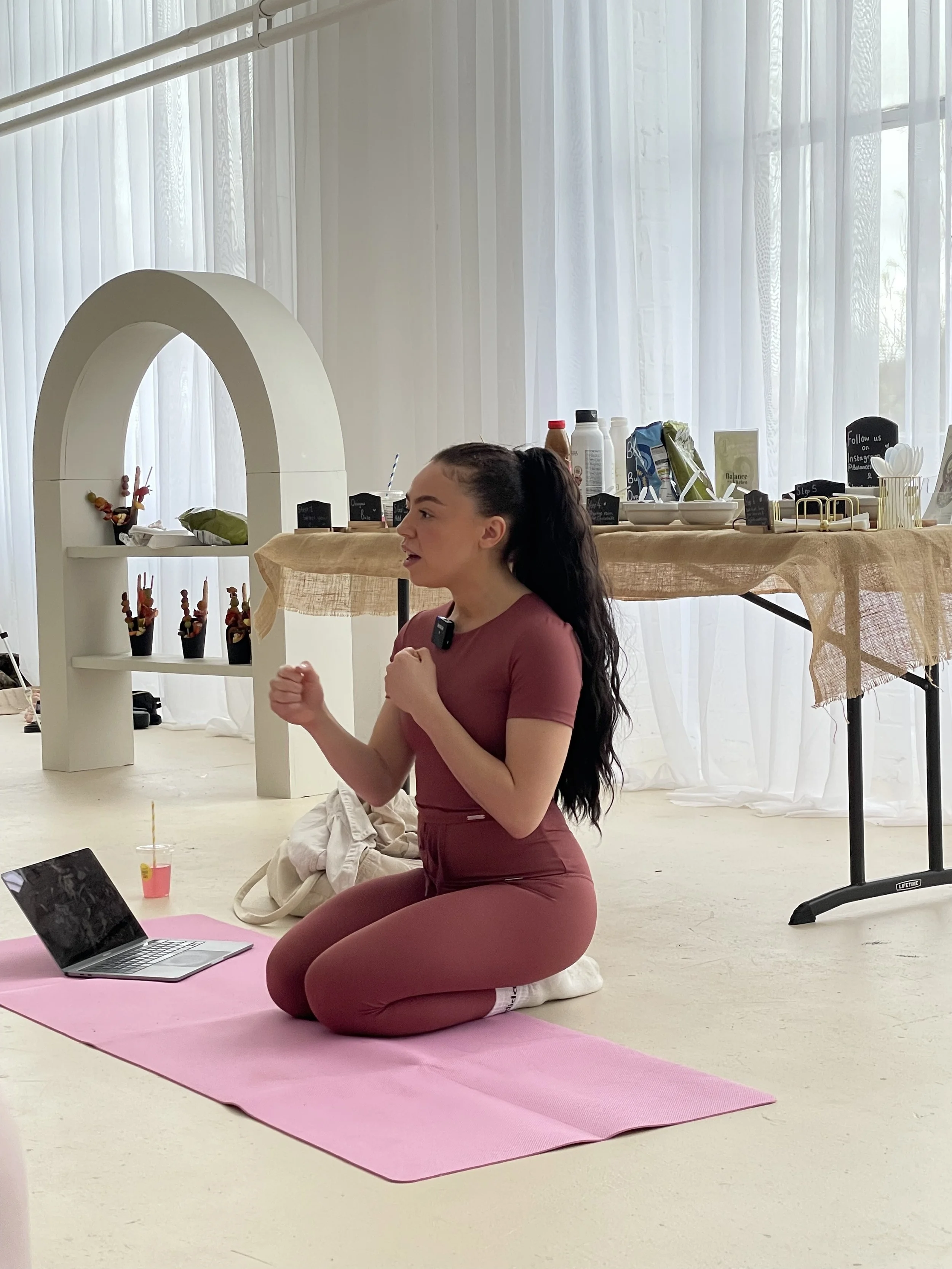 A young woman practicing yoga or meditation on a pink yoga mat in a bright room with white curtains. She is kneeling on her knees, with her hands held in a prayer position, wearing a maroon workout outfit.