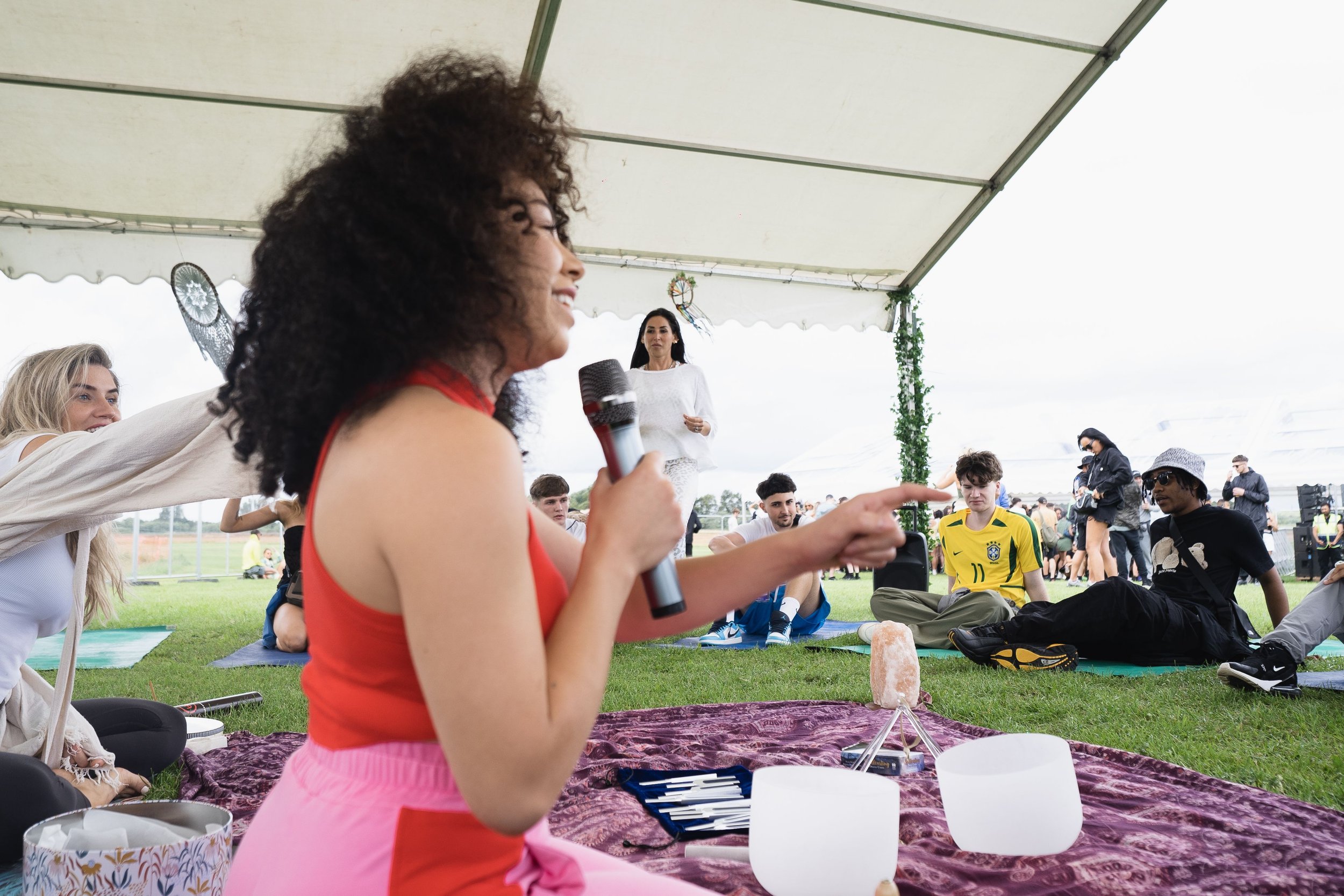 A woman with curly hair and a red top is speaking into a microphone in a tented outdoor setting with seated people and some standing in the background.