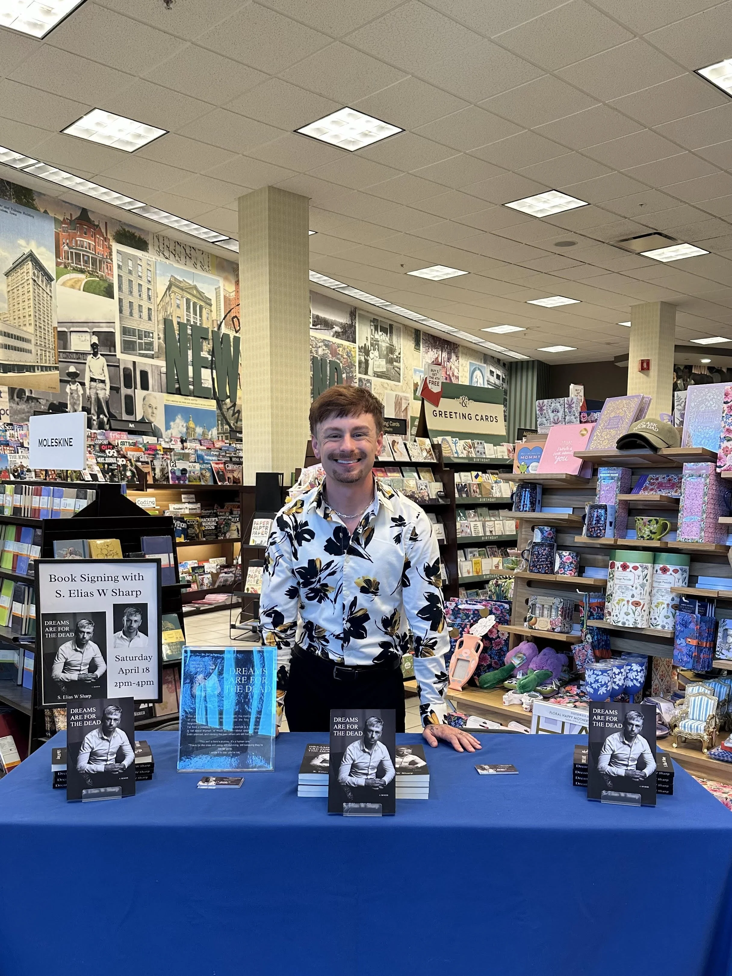 A man standing behind a table with books in a bookstore, promoting his book titled 'Dreams are for the Dead' during a book signing event. The sign indicates the event is scheduled for Saturday, April 18, from 2 pm to 4 pm.
