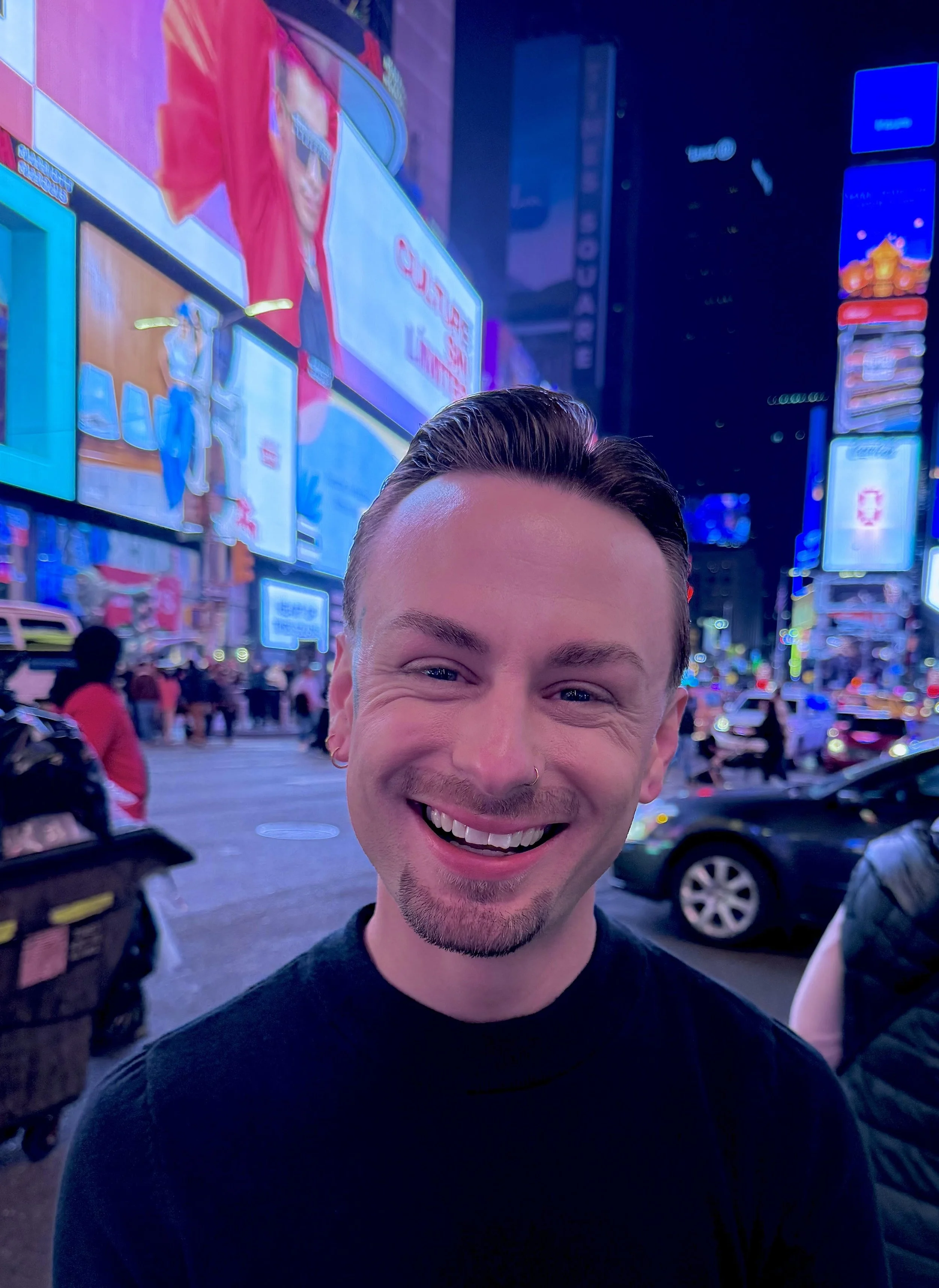 A smiling man with short dark hair, wearing a black shirt, standing on a brightly lit city street at night with illuminated billboards and electronic signs in an urban setting.