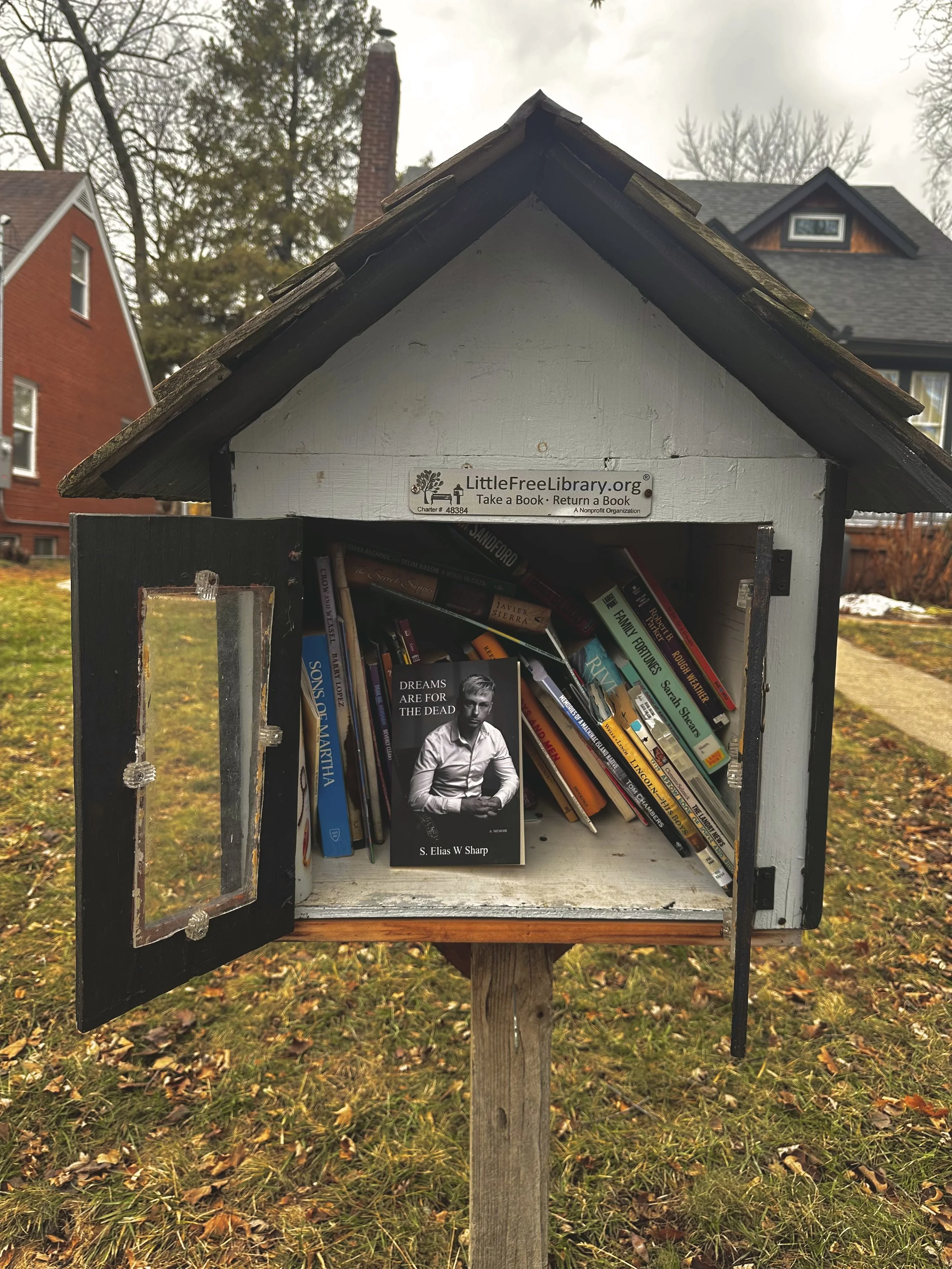 Small wooden library box on a post filled with books, including a black and white book cover titled "Dreams Are for the Dead" by S. Elias W Sharp, located in a yard with grass and trees in the background.