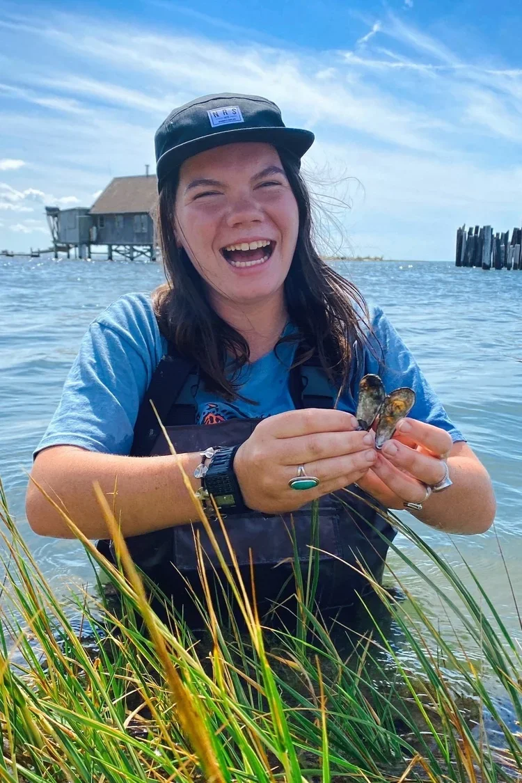 A smiling woman with a black cap, blue t-shirt, and a green ring on her finger, holding mussels she collected by the water, with a house on stilts and a dock in the background.