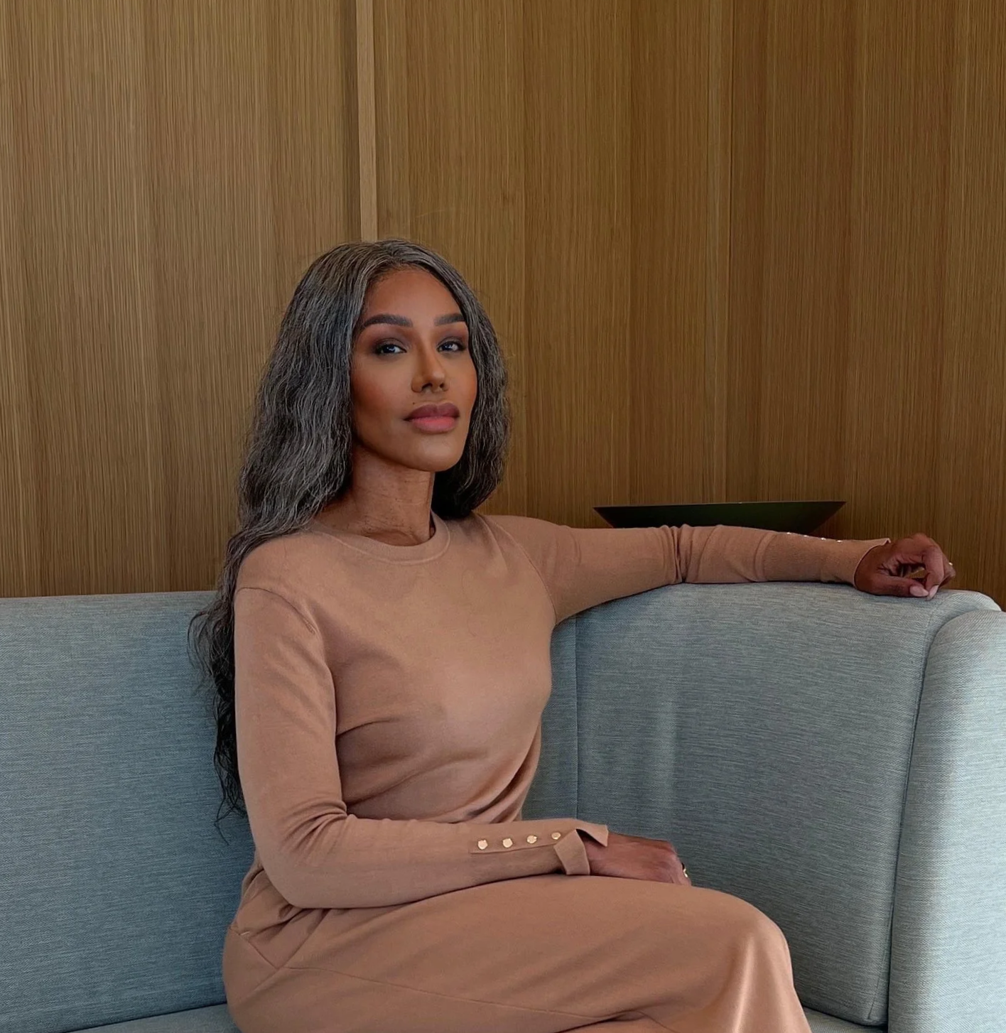 A woman with long gray hair and brown skin sitting on a gray sofa with a wooden wall background, wearing a beige long-sleeve top and matching pants. naida, diplomat, wellness coach, IIN, STRATEGIST, high performance. discipline, professional, expert,