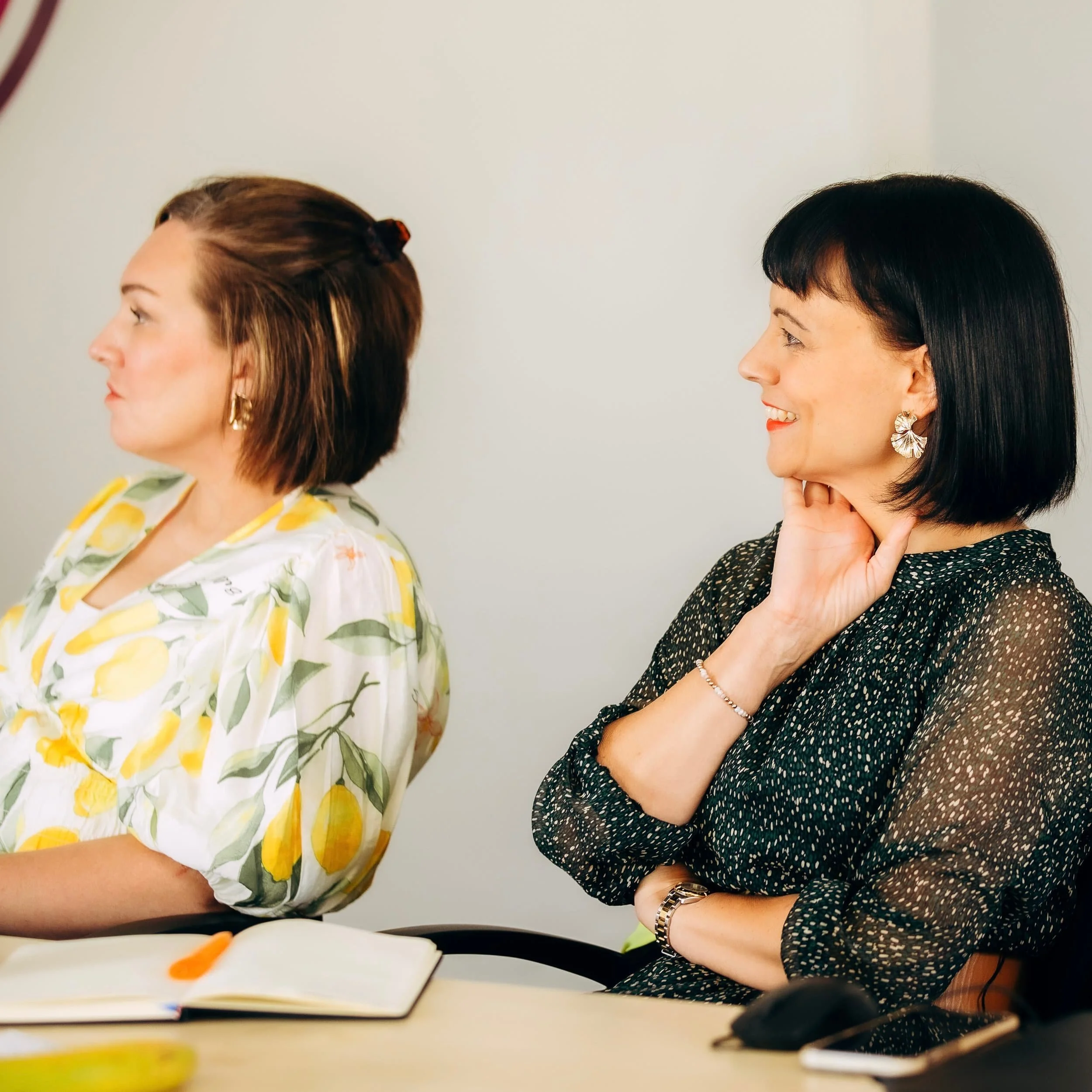 Two women sitting at a table, looking to their left, with open notebooks and a computer mouse on the table.