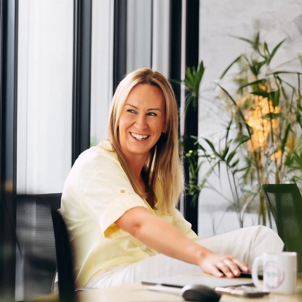 A woman with blonde hair sitting at a table in a modern office, smiling and looking to her right, with plants and large windows in the background.