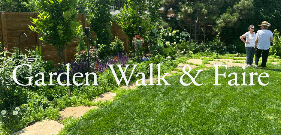 A lush garden with a stone pathway, green grass, and various plants and flowers. Two women are standing and talking in the background, with trees and a wooden fence surrounding the garden.