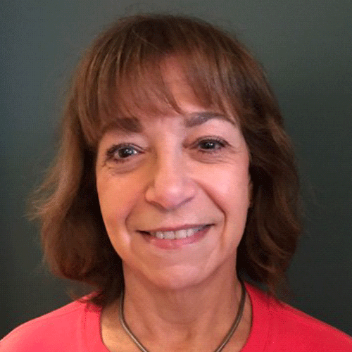 A woman with shoulder-length brown hair and bangs, smiling, wearing a red top and a silver necklace, against a dark background.