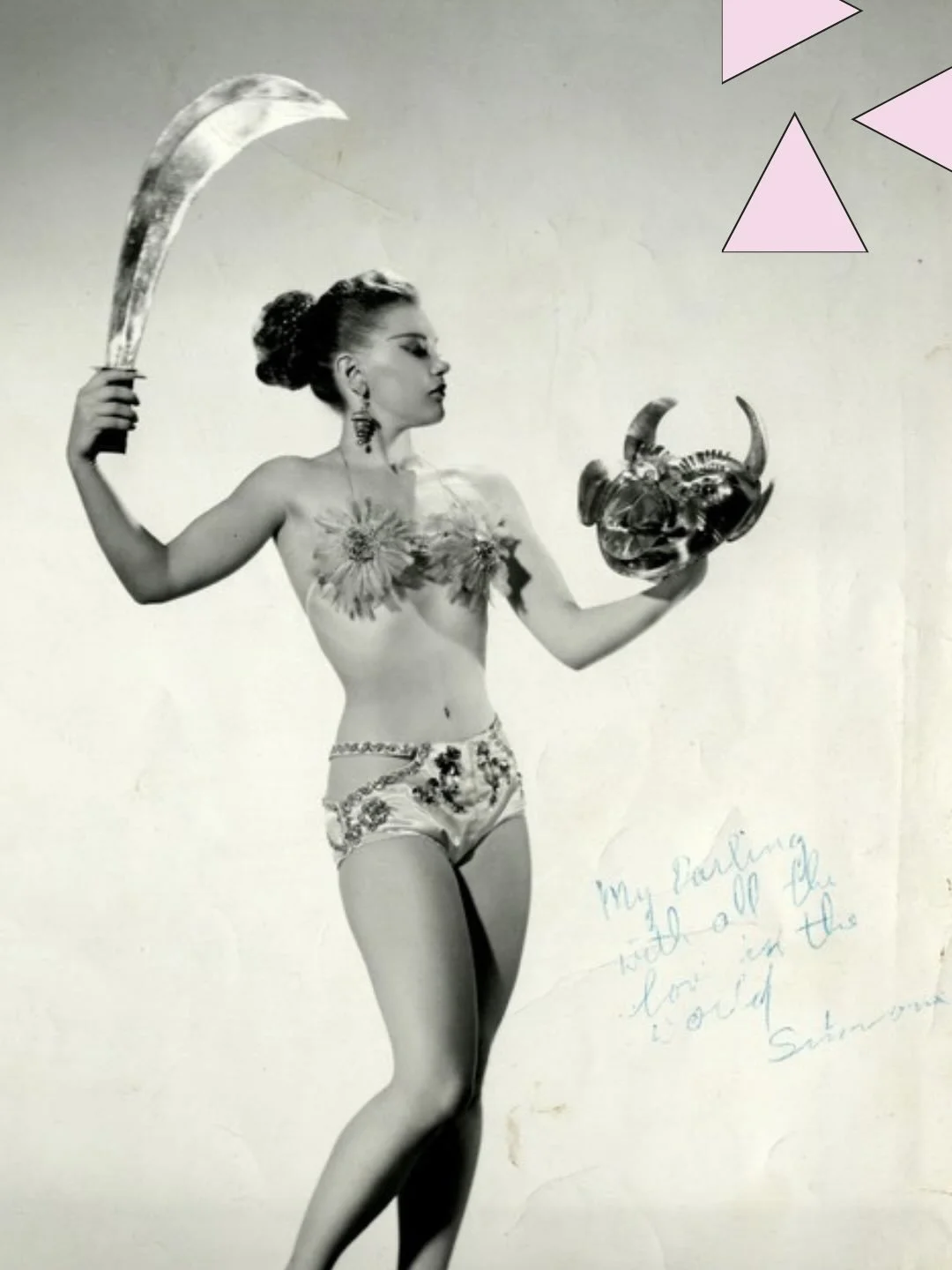 Mood.

Black-and-white portrait photograph of a woman theatrically posing with a sword and a mask in her hands, wearing a strappy, feathered bikini top and sequined satin panties with cut-aways at the hips, and spiral earrings. She has autographed th