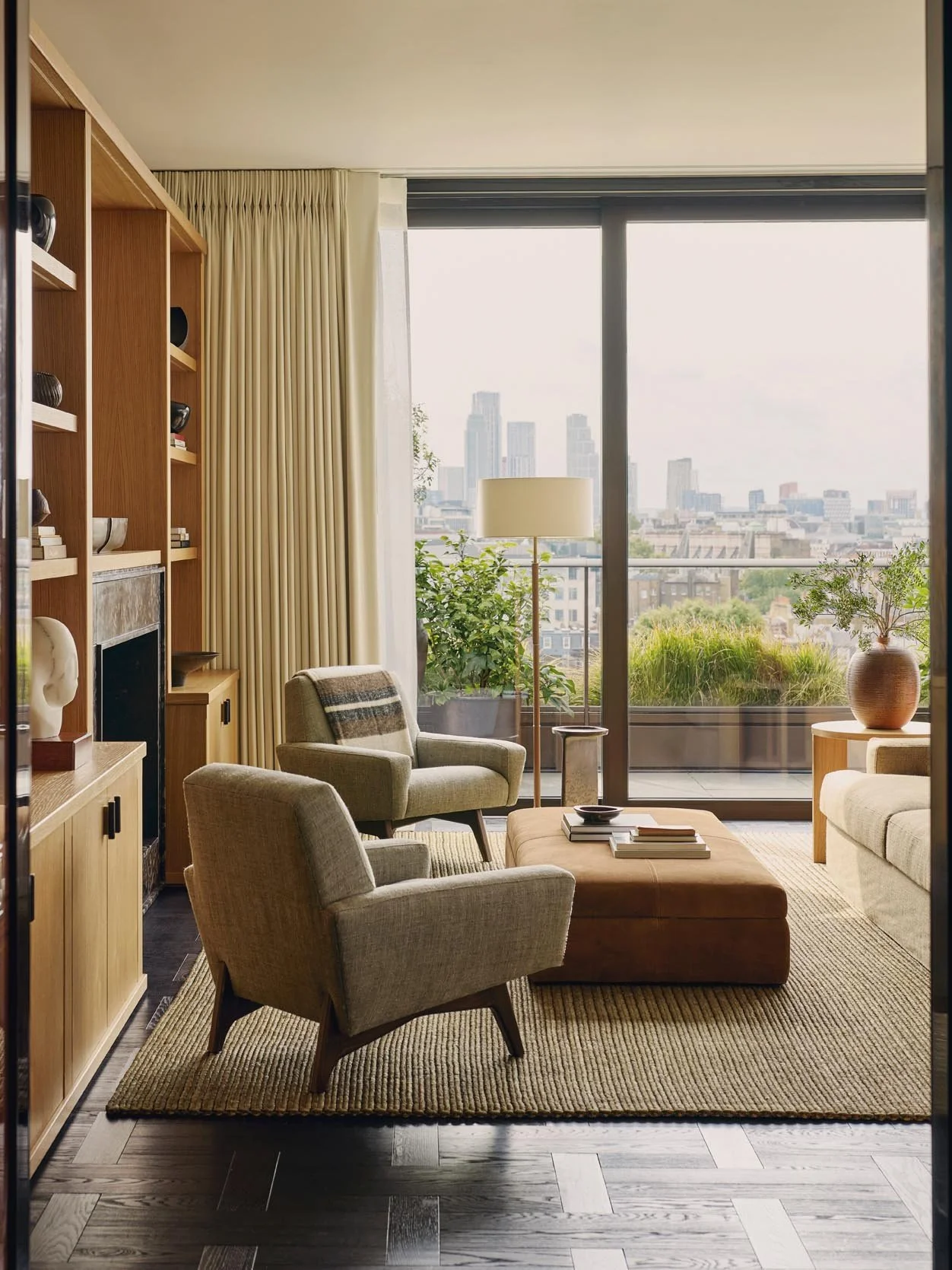 A sophisticated living room with beige armchairs and a brown sofa, a suede ottoman with books, potted plants, a tall floor lamp, and large glass sliding doors overlooking the city at The Peninsula Hotel Residences.