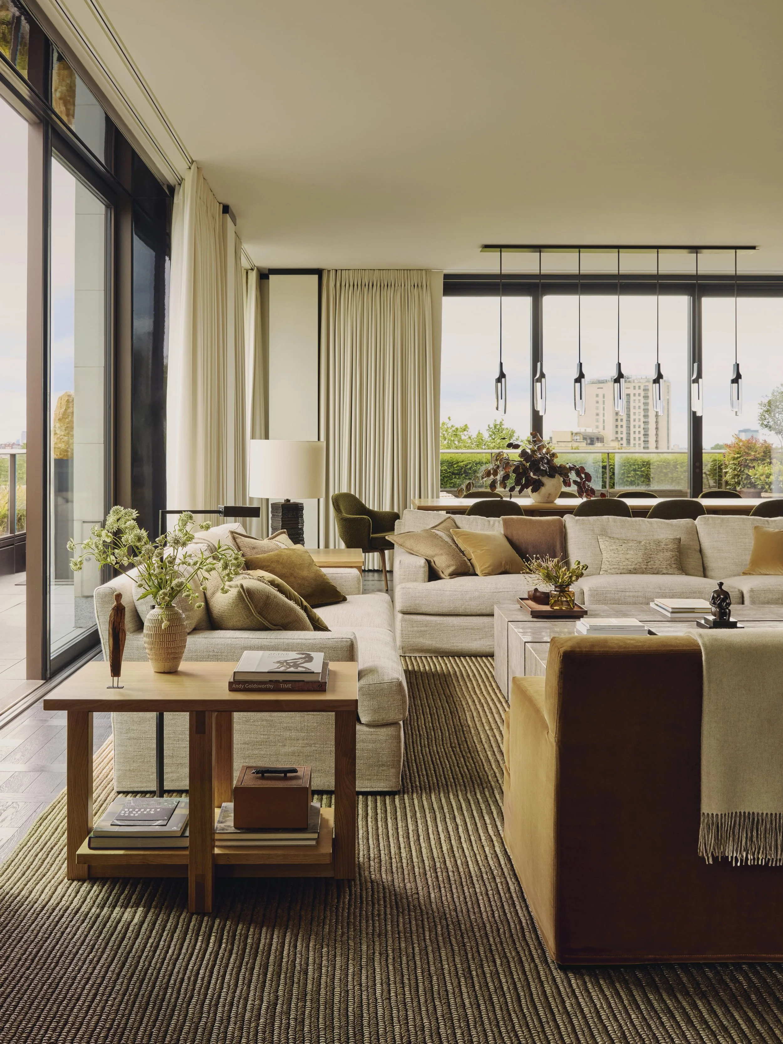 Modern and warm living room with large windows, beige sofas with pillows, a wooden side table with books and a vase, a natural weave carpet. coffee table with decorative items, and a view of city buildings outside. In the Peninsula Hotel Residences.