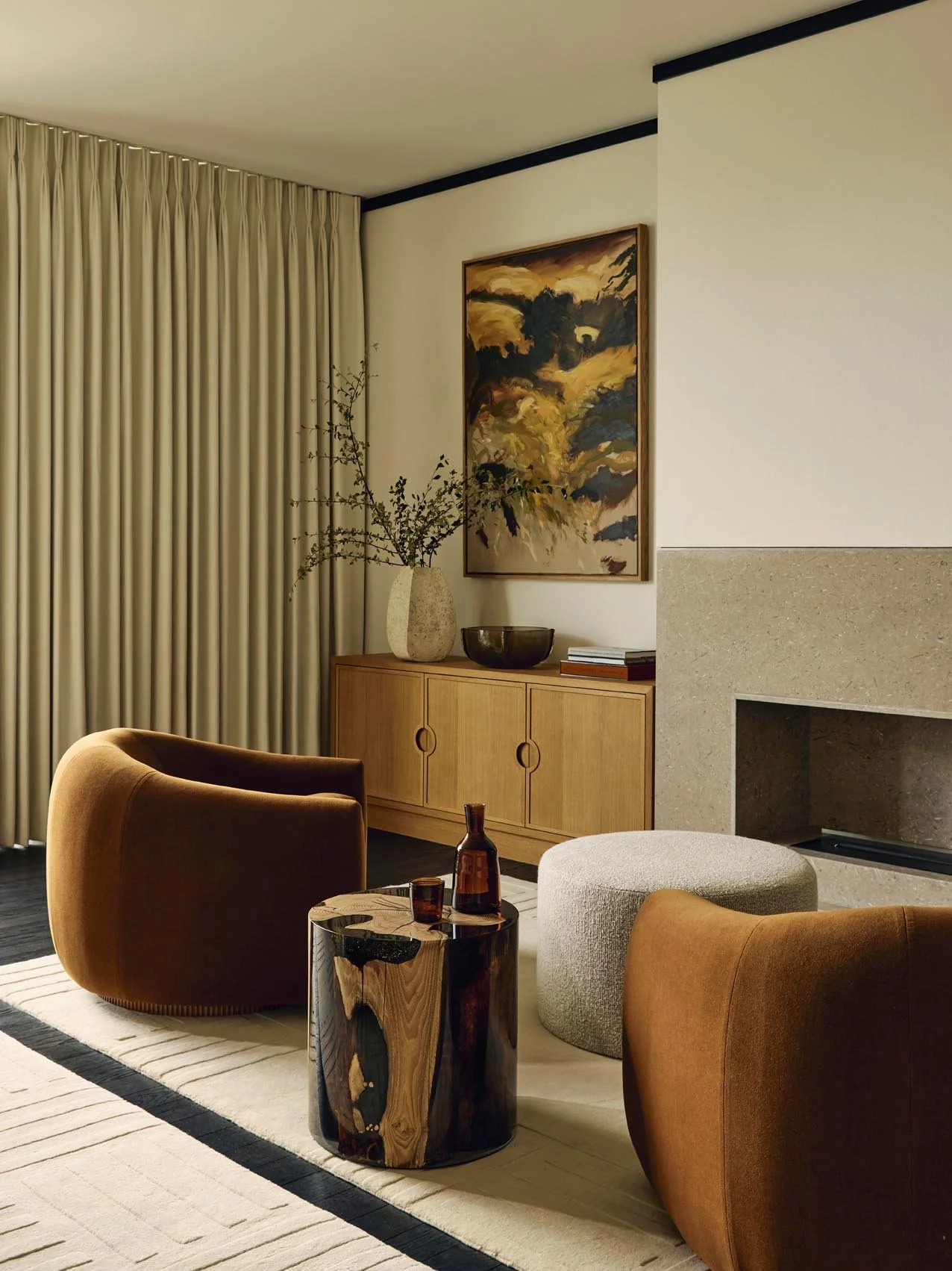 Cozy bedroom lounge at The Peninsula Hotel Residences featuring two brown modern armchairs, a marble fireplace, a light beige pouf, and a wooden sideboard curated with books, a ceramic vase, and abstract artwork, complemented by beige curtains.