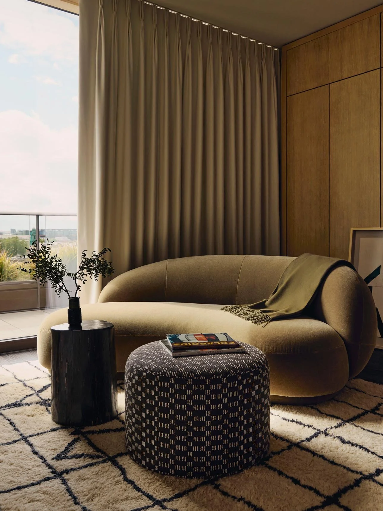 Modern bedroom lounge at The Peninsula Hotel Residences with green velvet sofa, patterned ottoman, black side table with greenery, beige curtains, moroccan rug and urban city views.