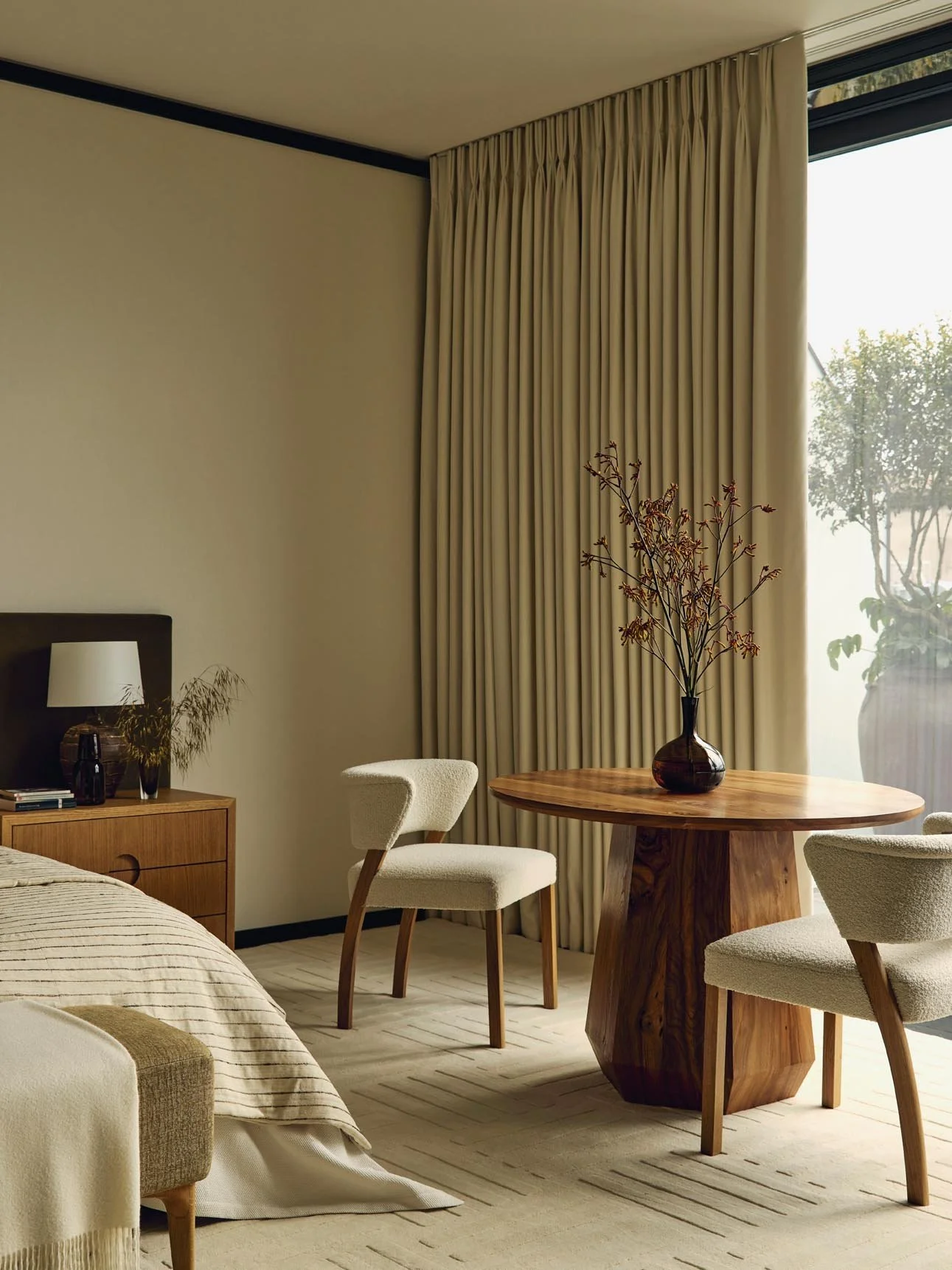 A serene bedroom corner featuring a bed with a wooden nightstand, two beige upholstered chairs, a round wooden table with a vase of branches, beige curtains, and a large window overlooking a potted plant at The Peninsula Hotel Residences.