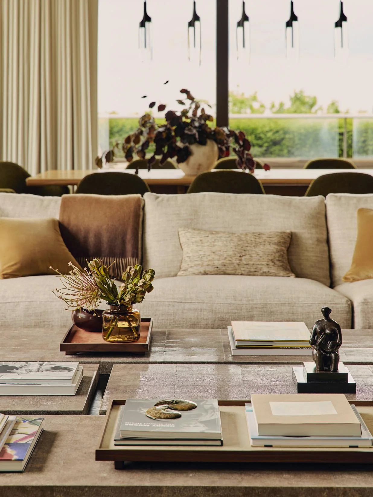 A sophisticated living room with a sofa and decorative pillows, a coffee table with books, a small sculpture, and a vase of flowers, overlooking a dining area and a window with lush greenery at The Peninsula Hotel Residences.