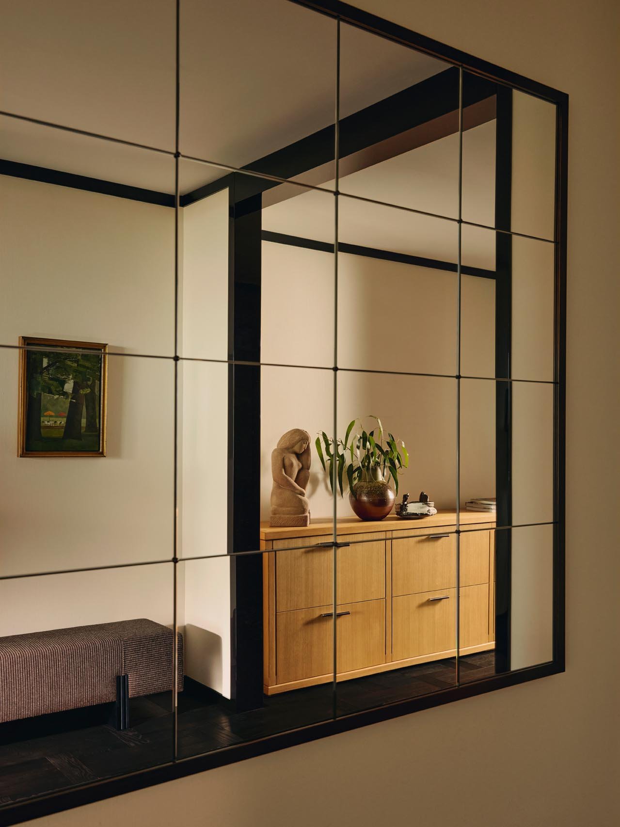 Decorative Mirror in the entrance of an apartment at The Peninsula Hotel Residences, with a grid-pattern mirror reflecting a refined interior with a wooden sideboard, sculpture a ceramic potted plant, and carefully arranged books.
