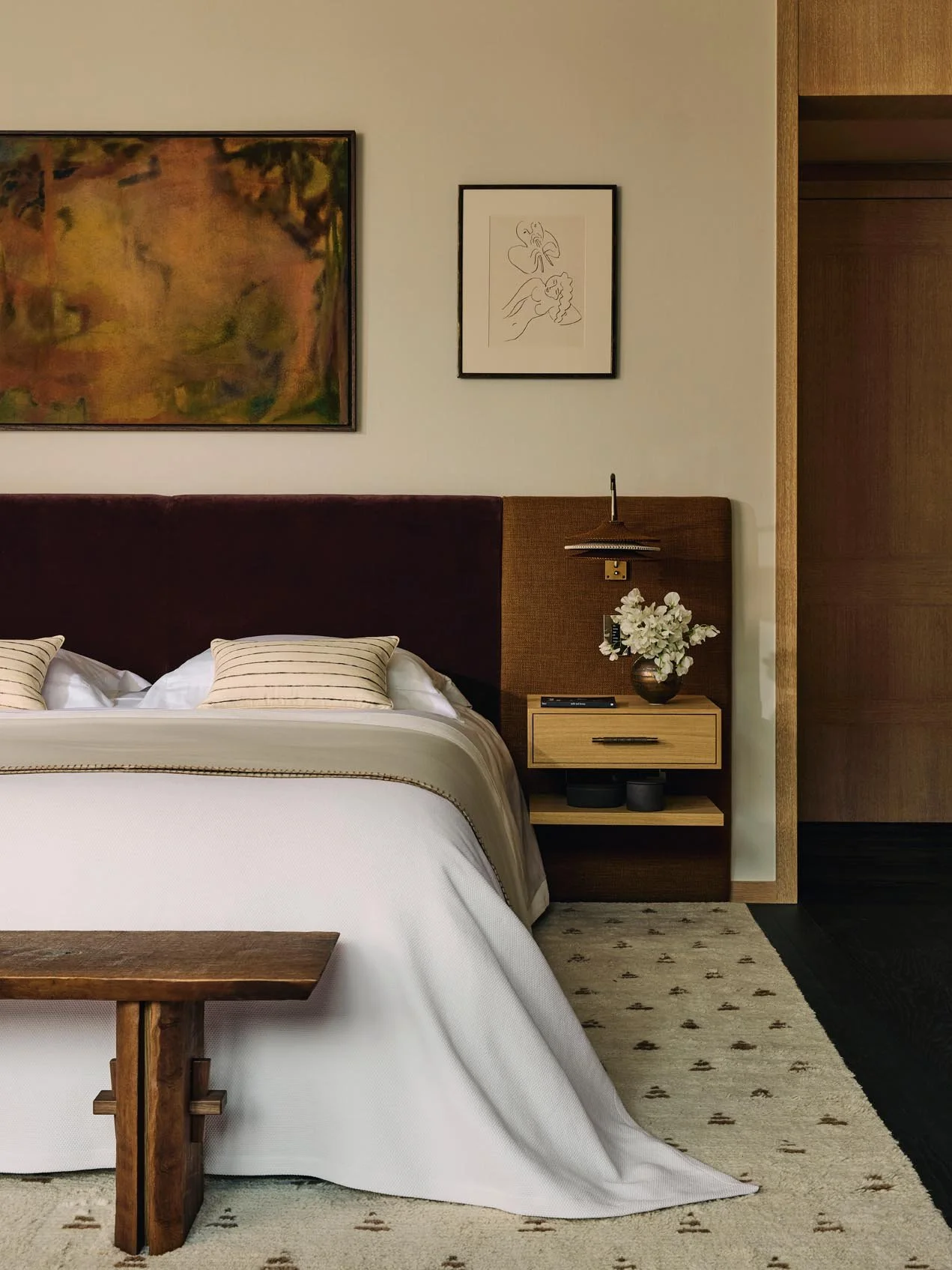 Luxury bedroom at The Peninsula Hotel Residences with a dark burnt orange and brown headboard bed, white bedding, striped pillows, wooden bench, nightstand with vase, and curated artwork, set over a beige and burnt orange patterned rug.