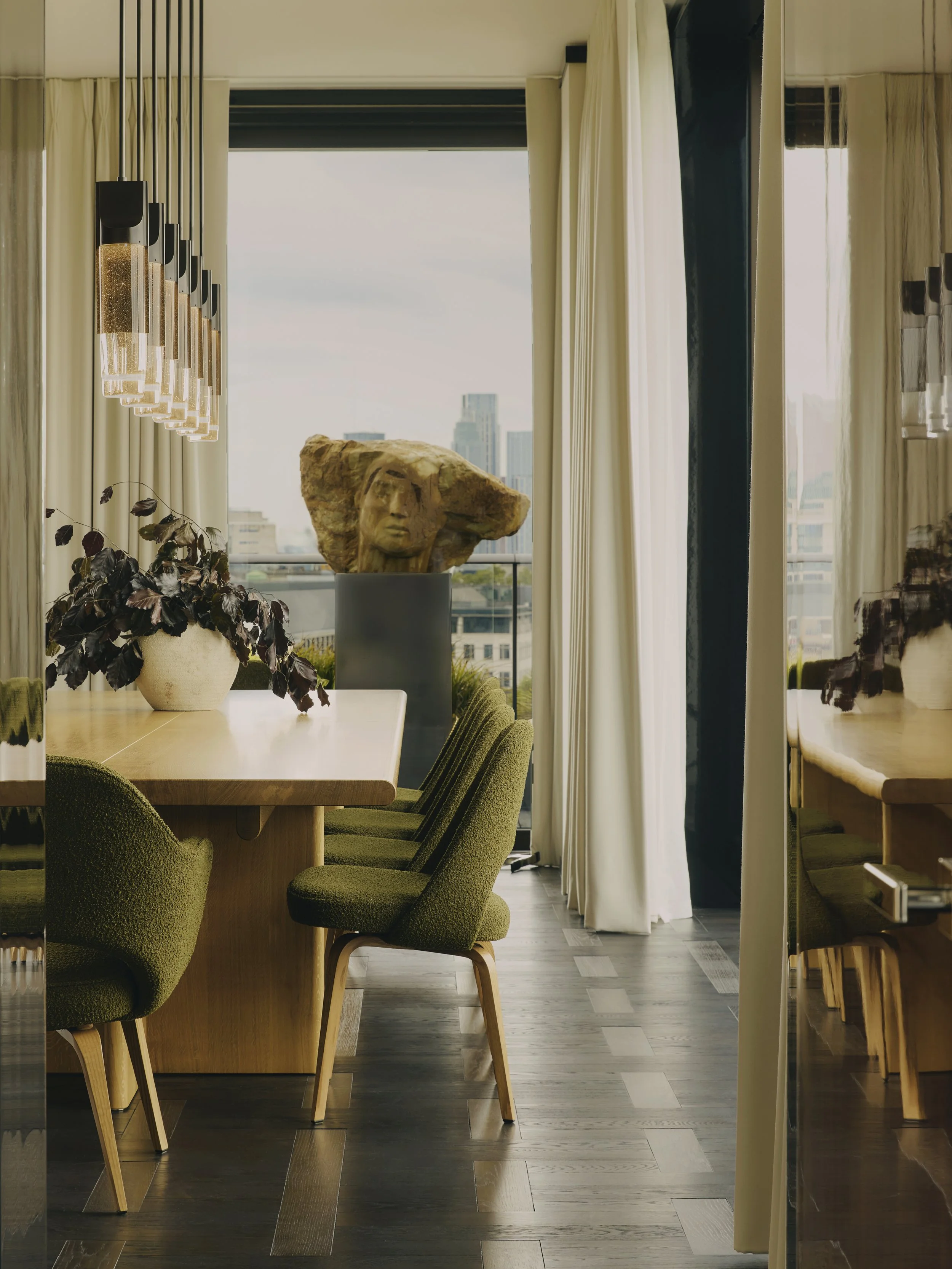 Elegant Dining Room at The Peninsula Hotel Residences, a sophisticated space featuring a wooden dining table, green upholstered chairs, and a stone face sculpture on a pedestal, bathed in natural light from large windows with city views.