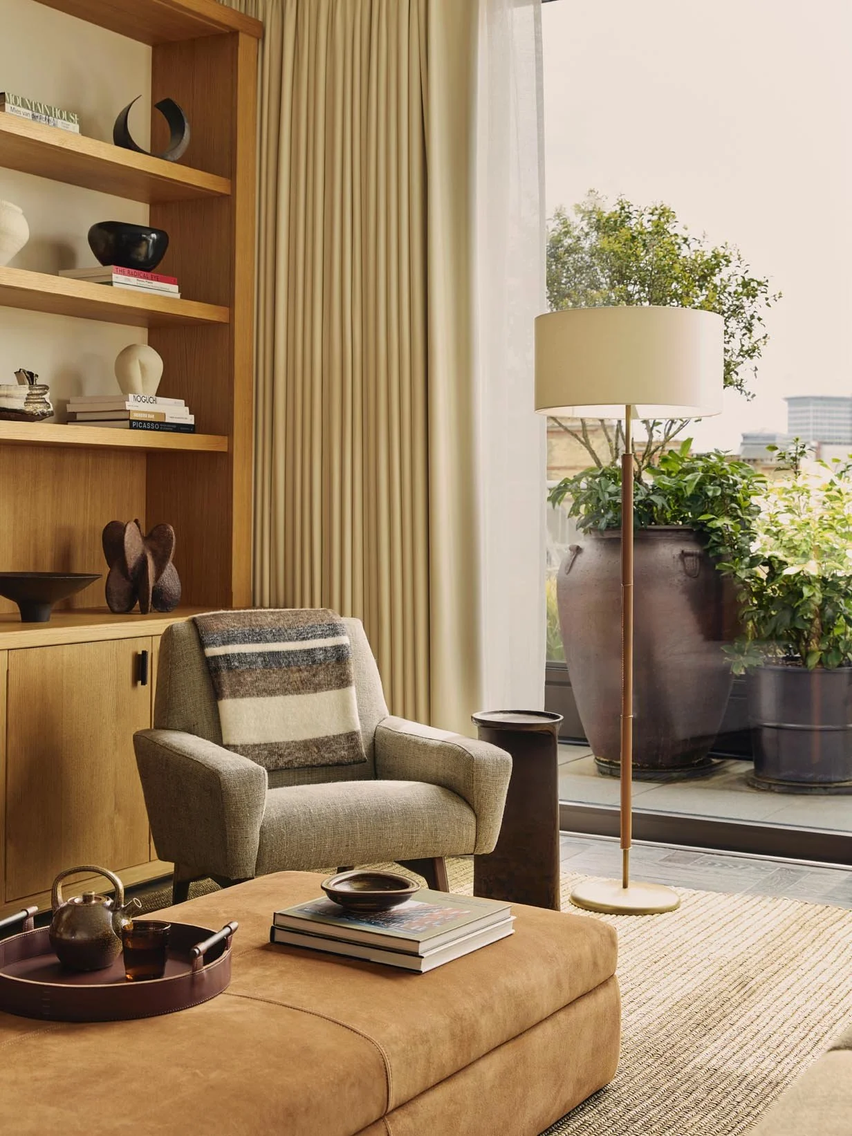 Luxury living room at The Peninsula Hotel Residences featuring beige armchairs, a brown sofa, a suede ottoman with books, potted plants, a tall floor lamp, and large glass sliding doors with city views.