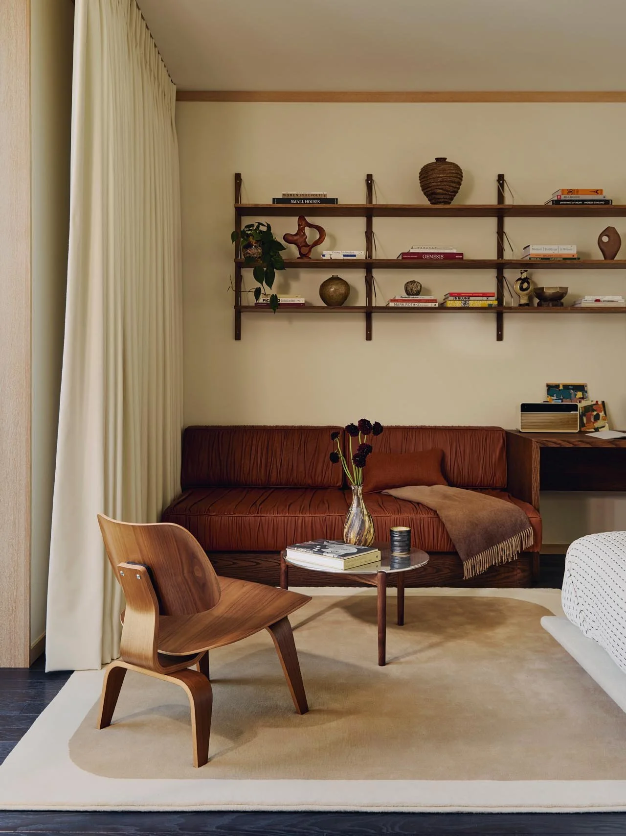 A bedroom lounge with beige curtains, a leather sofa with throw and book, an Eames wooden chair, Scandinavian wall-mounted shelves with books and objets, and a beige rug, styled in a vintage-meets-modern mix at The Peninsula Hotel Residences.