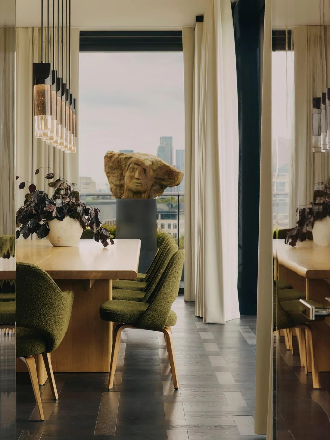 Luxury modern dining area at The Peninsula Hotel Residences featuring a wooden table, green upholstered chairs, a cast bronze and glass pendant, and a large stone sculpture of a woman’s face, with cityscape views through the window.