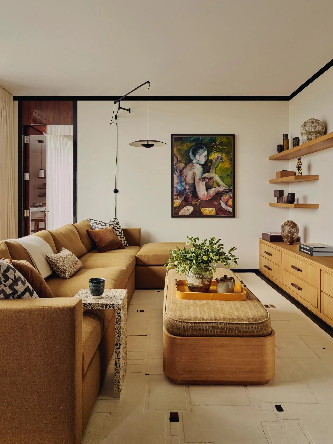 A warm and stylish TV room with a mustard corner sofa, patterned cushions, wooden ottoman with tray, side table, artwork, and curated shelving at The Peninsula Hotel Residences.