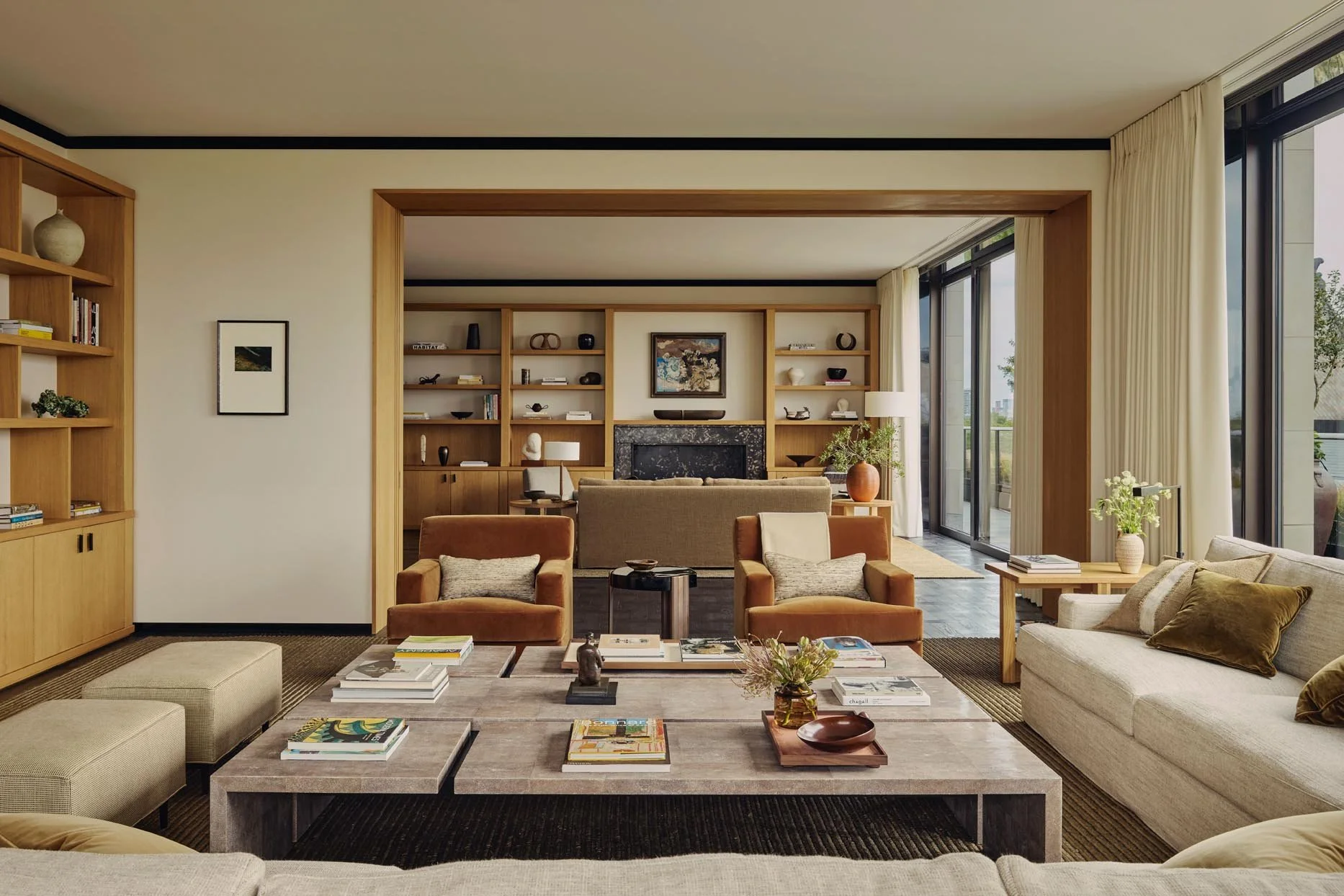A modern, warm living room with beige and brown furniture in earthy tones, wooden bookshelves, large glass windows with city views, and a curated collection of art and decorative objets at The Peninsula Hotel Residences.