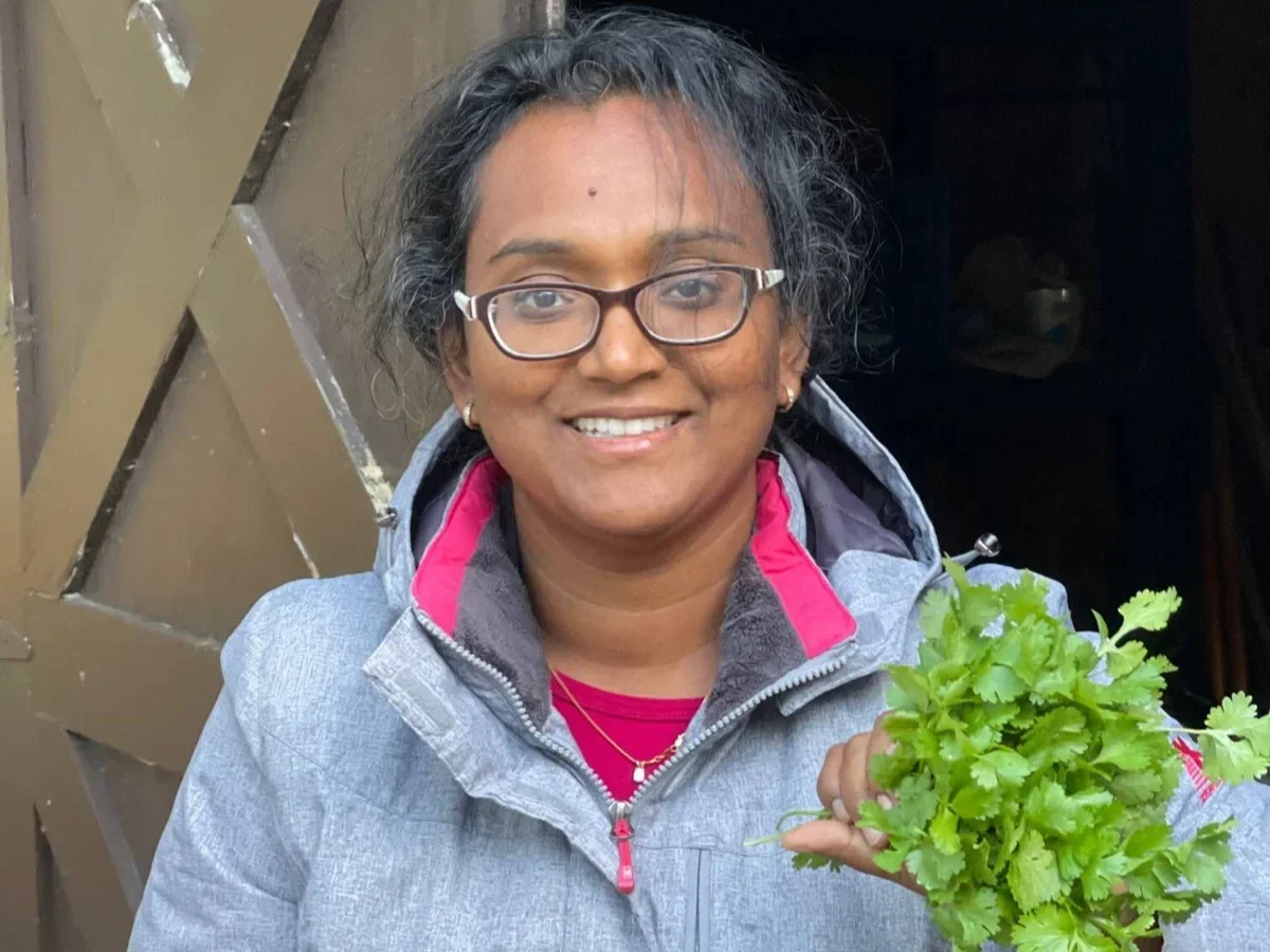 A woman with glasses smiling and holding a bunch of cilantro or parsley in front of a wooden wall or door.