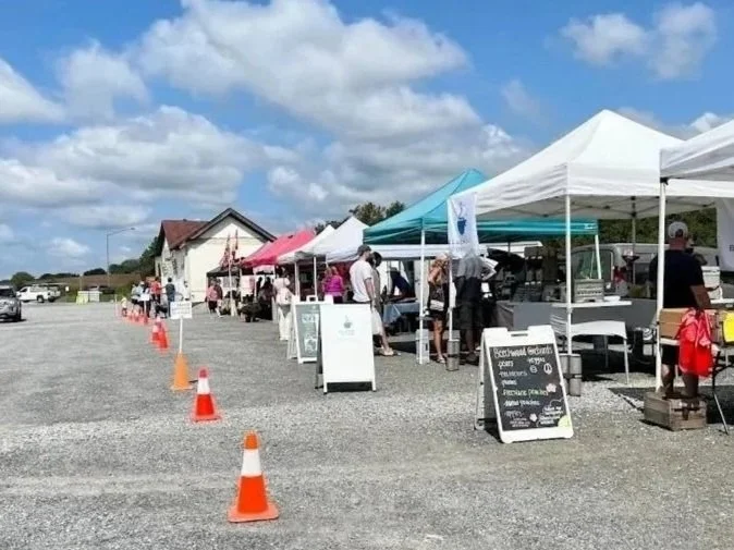 Gilbert's Corner farmers market Aldie, VA with white and colored tents and people shopping, orange traffic cones along a gravel lot, under a partly cloudy sky.