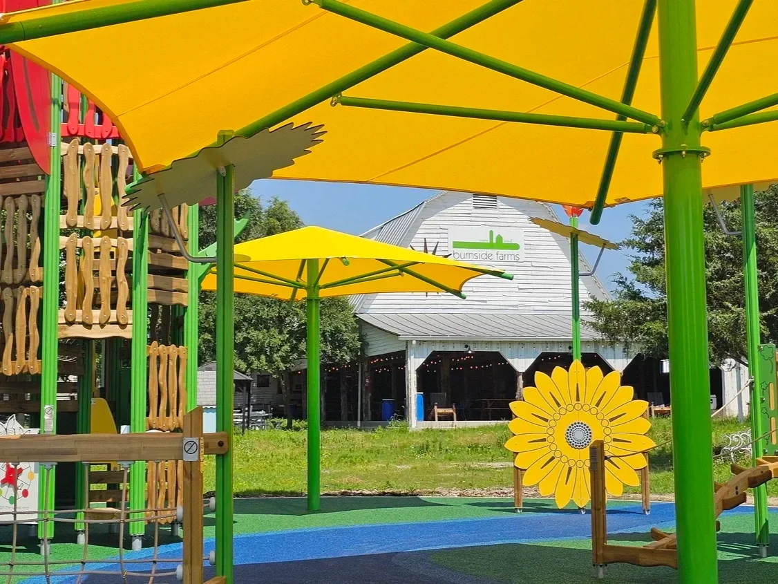 Colorful outdoor playground with bright yellow umbrellas, wooden play structures, and a white barn in the background with a green 'Burnside Farms' sign.