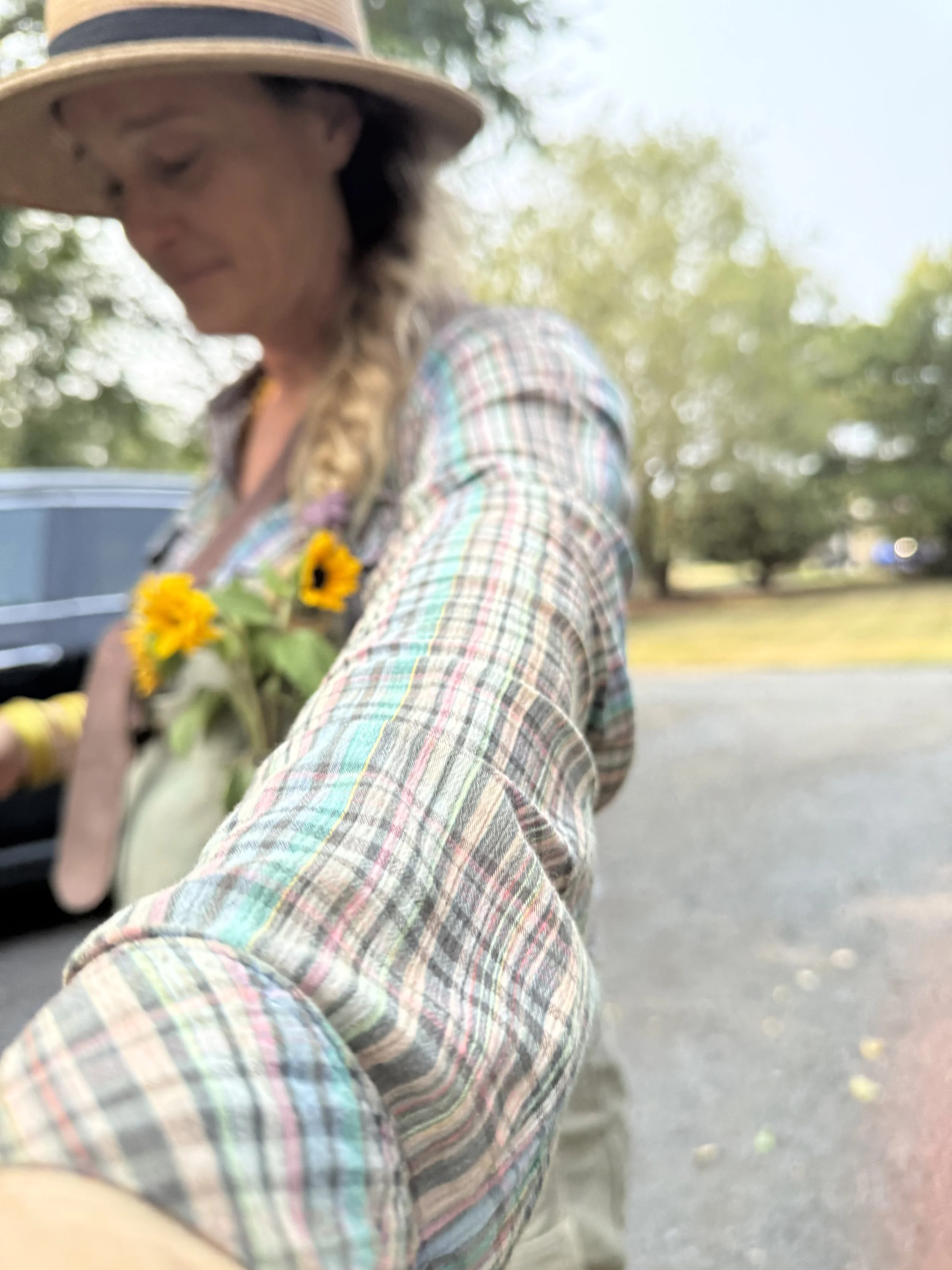 Woman wearing a wide-brimmed hat, plaid shirt, and a flower pin, reaching out towards the camera outdoors with trees and a car in the background.