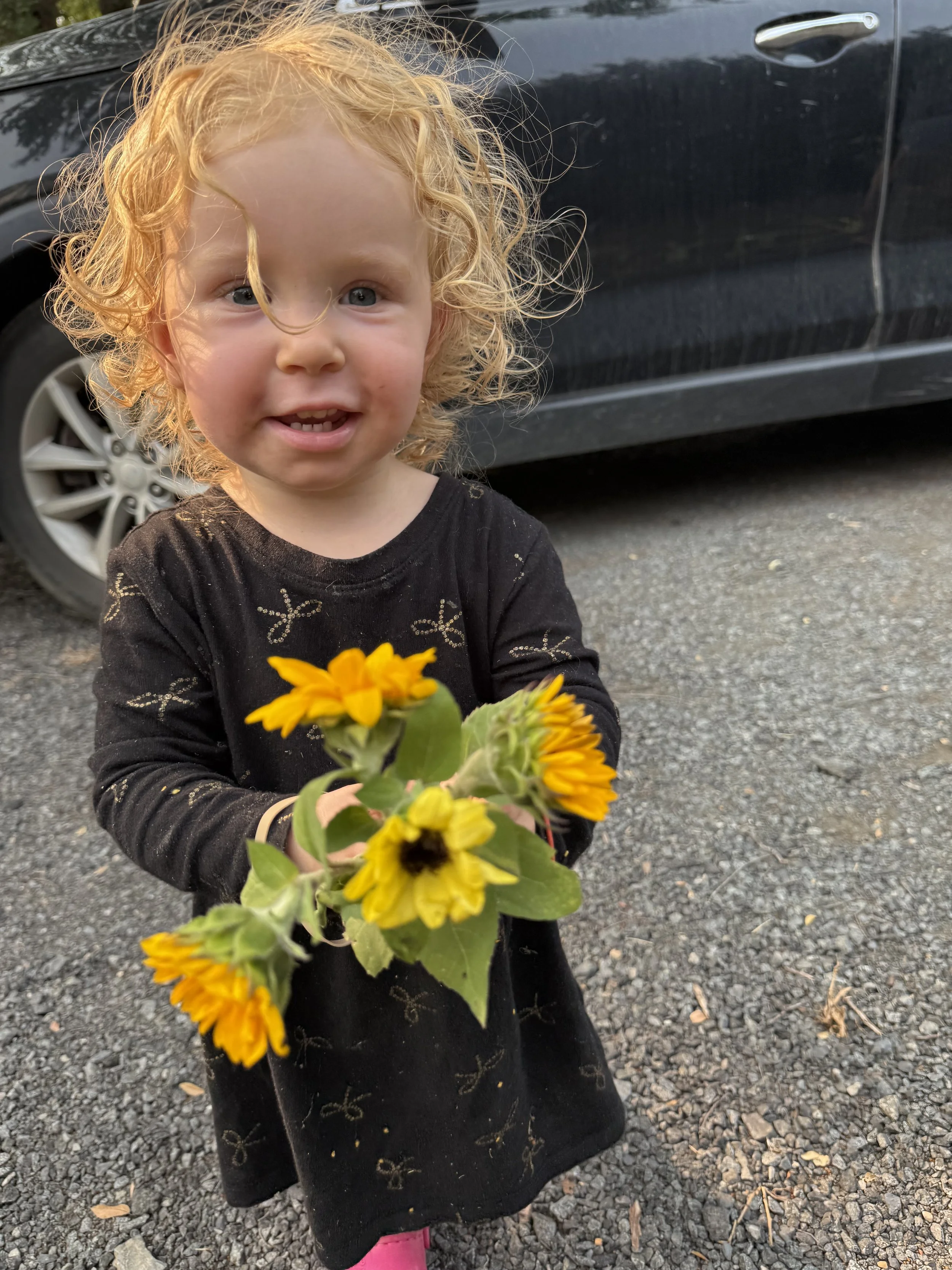 A young girl with curly blonde hair and a black dress standing outdoors, holding a bunch of yellow flowers, smiling at the camera.