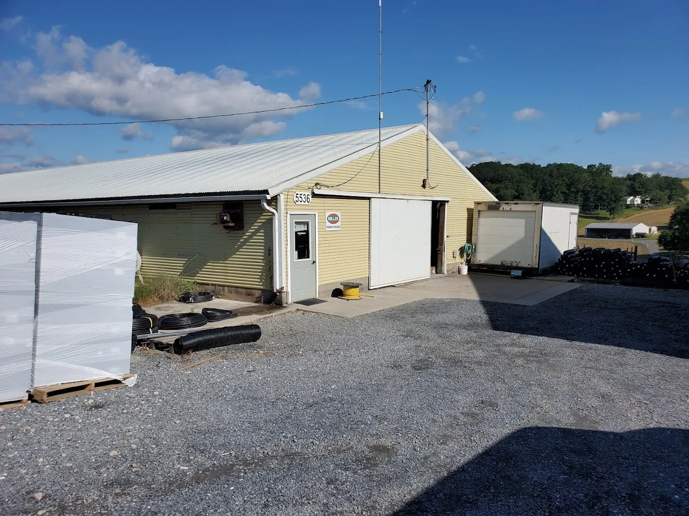 A yellow metal building with a white roof, a door, and a large sliding door. There is a trailer and stacks of black pipes outside on a gravel lot, with a blue sky and some clouds in the background.