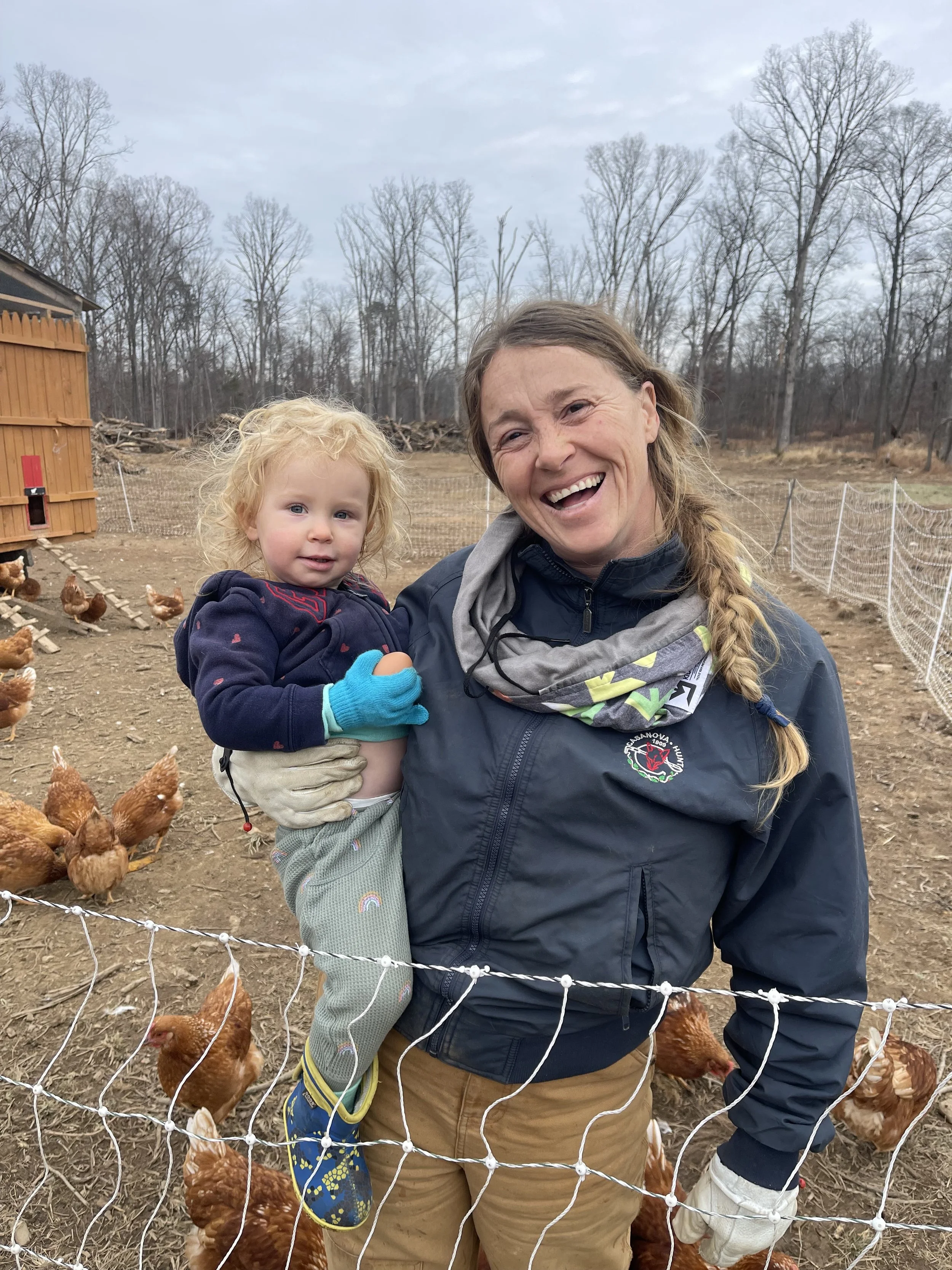 A woman and a young girl at a farm with chickens, woman smiling, girl wearing cycled gloves, barn in the background.