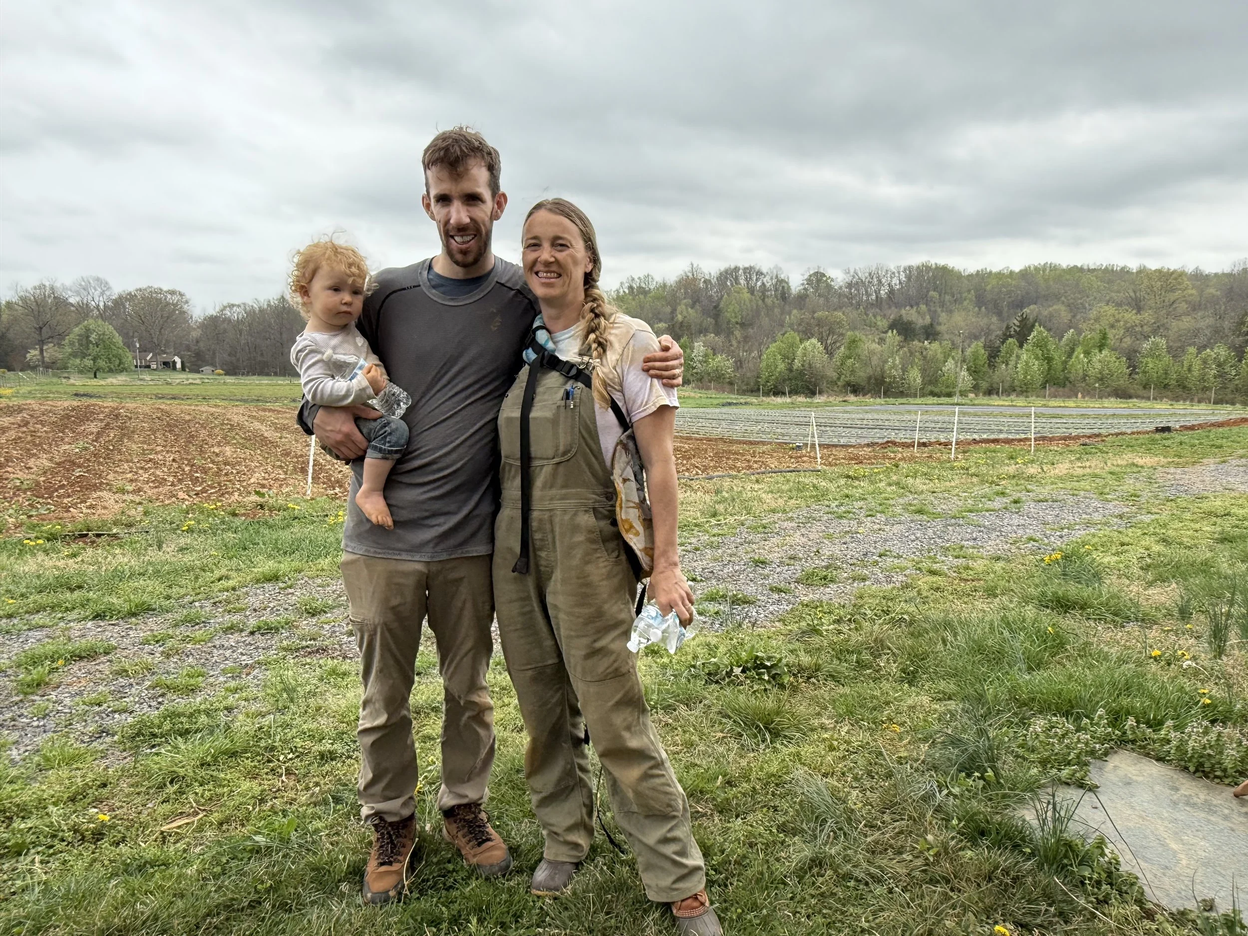 A happy family of three standing outdoors on a cloudy day, with a farm and green trees in the background. The man is holding a child, and the woman is hugging the man. They are all smiling.