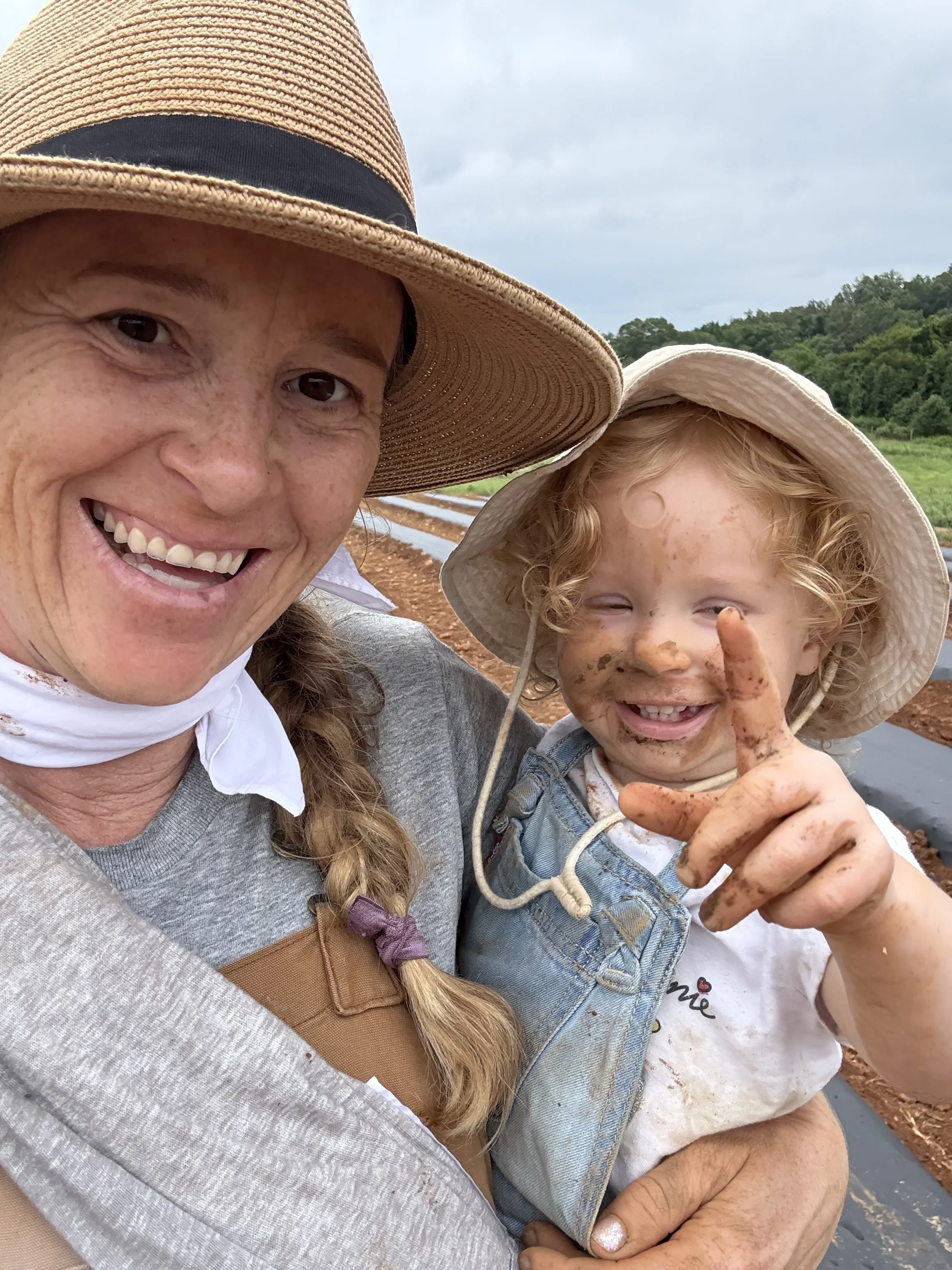 A woman and a young girl outdoors in a field, smiling happily. The girl has muddy face and hands, wearing a beige sun hat and overalls, and the woman is wearing a wide-brimmed hat and a gray shirt.