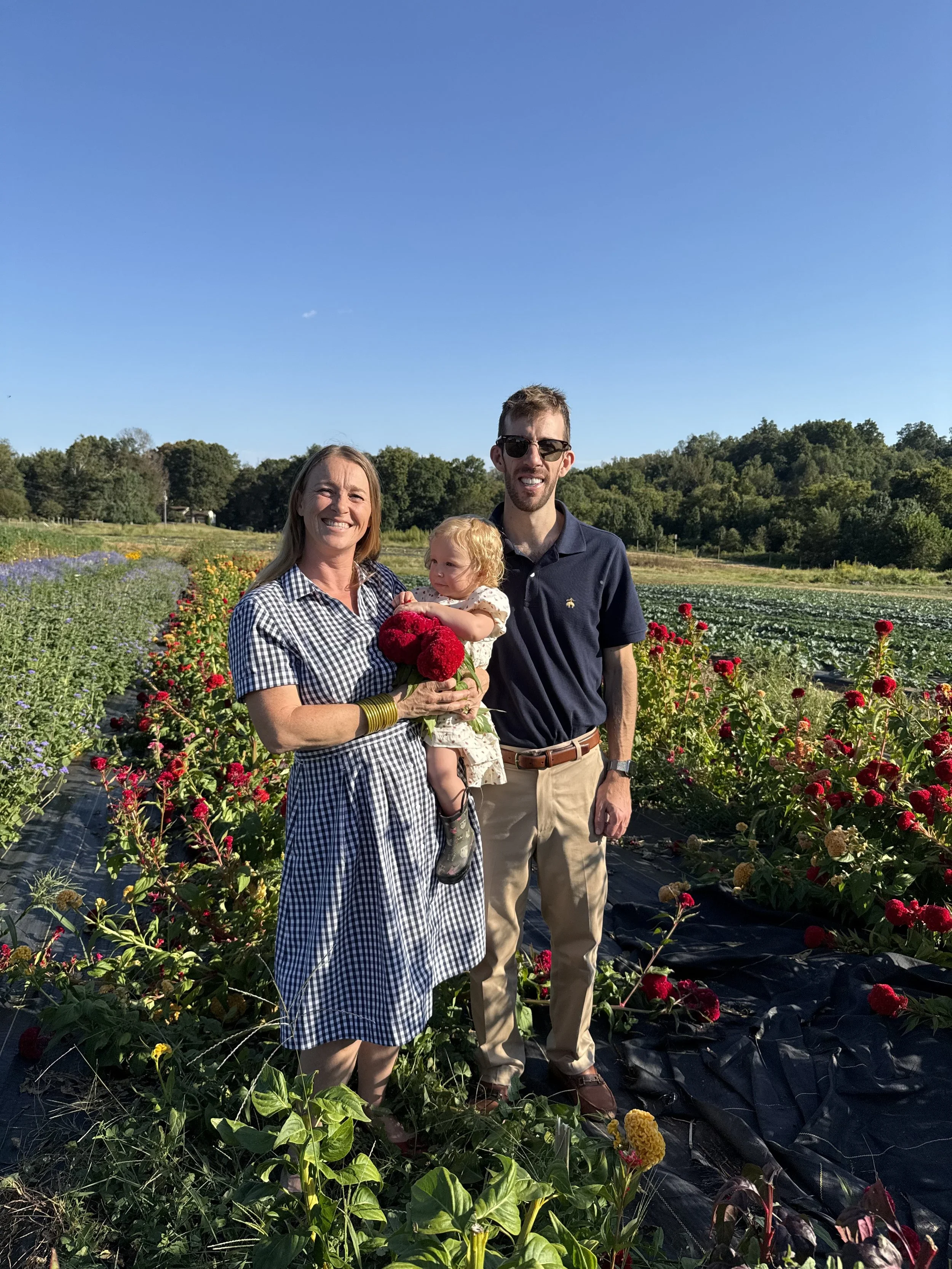 A family of three standing in a flower field, smiling and holding freshly picked flowers on a sunny day.