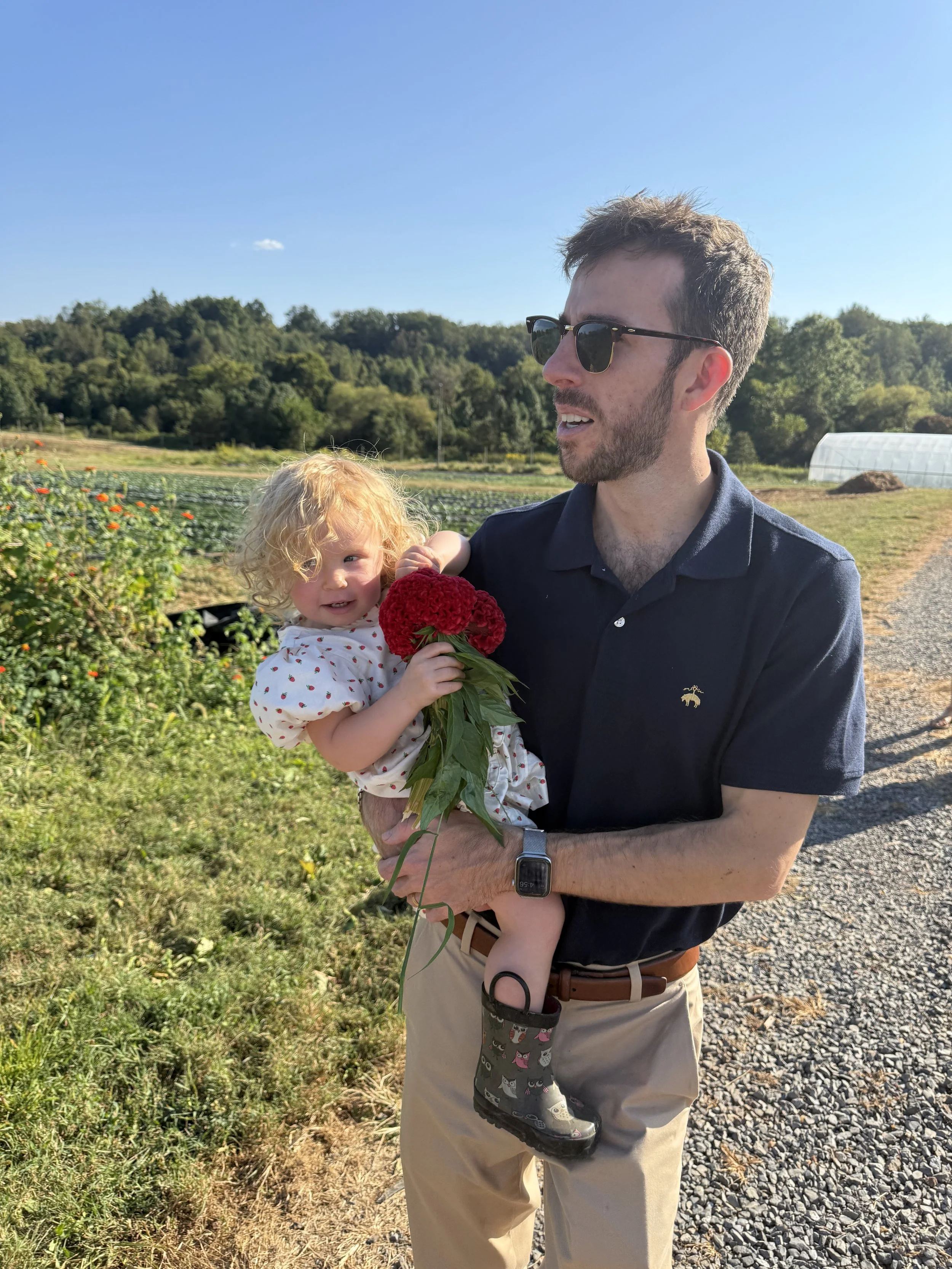 A man wearing sunglasses and a navy blue polo shirt holding a young girl with blonde curly hair, a white dress with red polka dots, and rain boots with a cat pattern, outdoors in a farm or garden setting with green fields and blue sky.