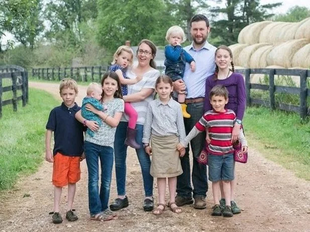 Family of nine standing on a dirt path at a farm with green trees and hay bales in the background, holding hands and smiling.
